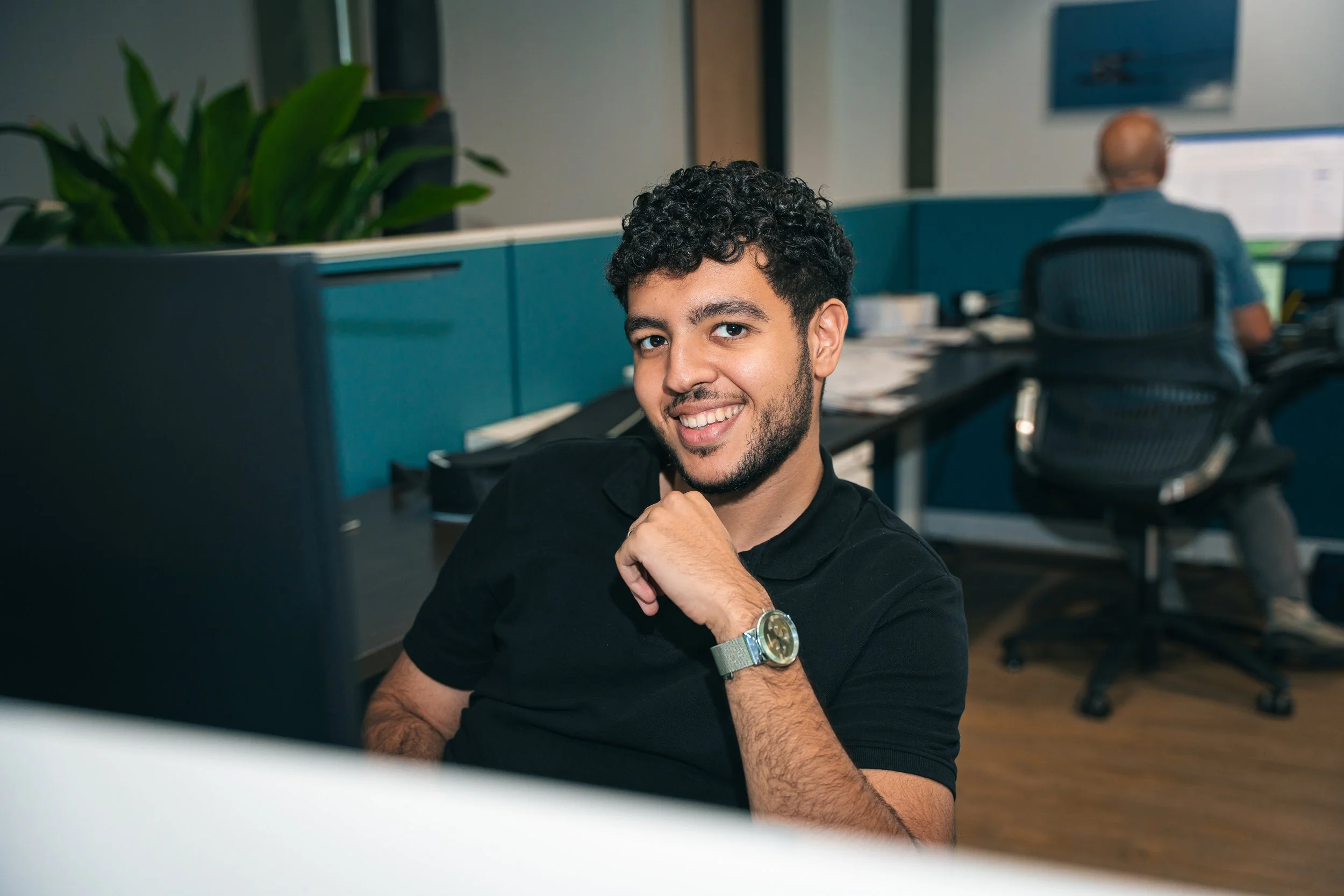 A young man with curly hair and a beard smiling at the camera, sitting at a desk in an office environment.