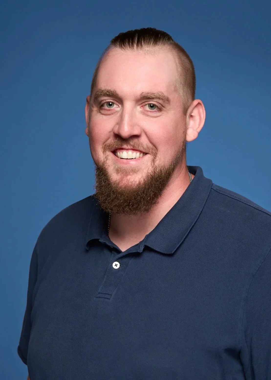 A young man with a beard and short hair wearing a navy blue polo shirt, smiling against a blue background.