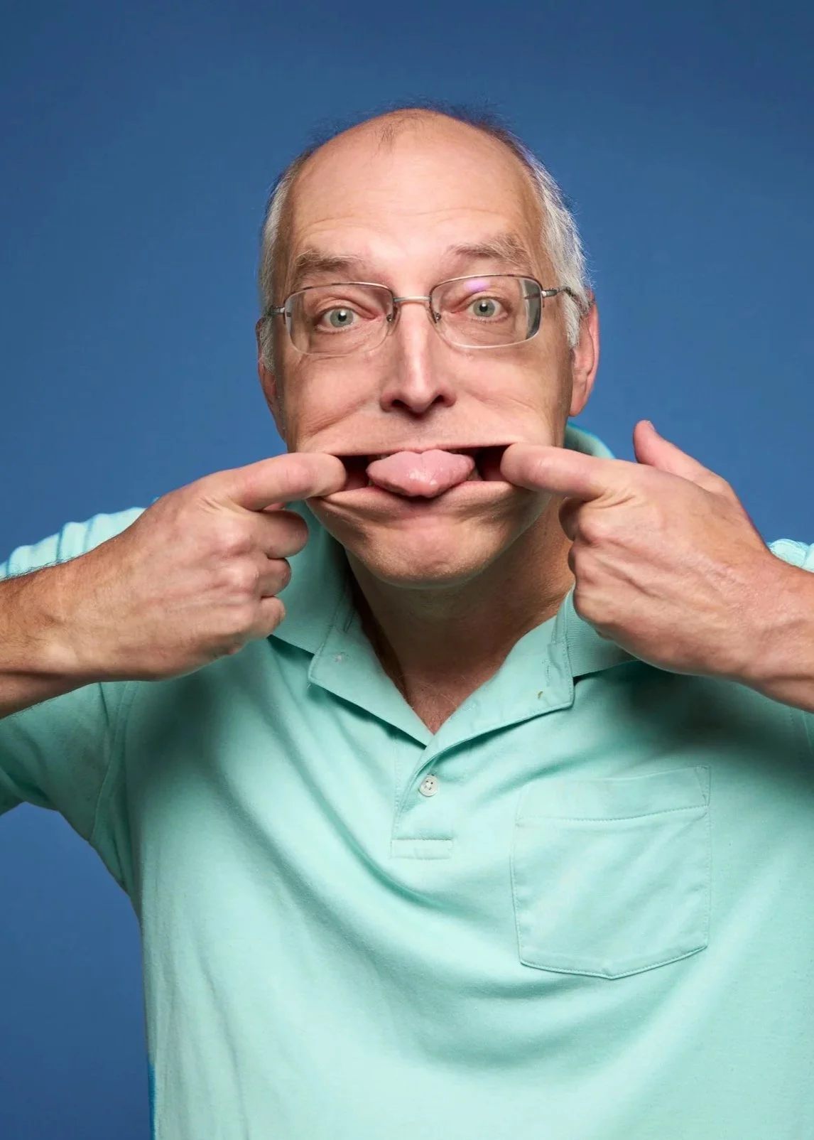 A middle-aged man with glasses making a funny face by pulling the corners of his mouth with his fingers and sticking out his tongue.