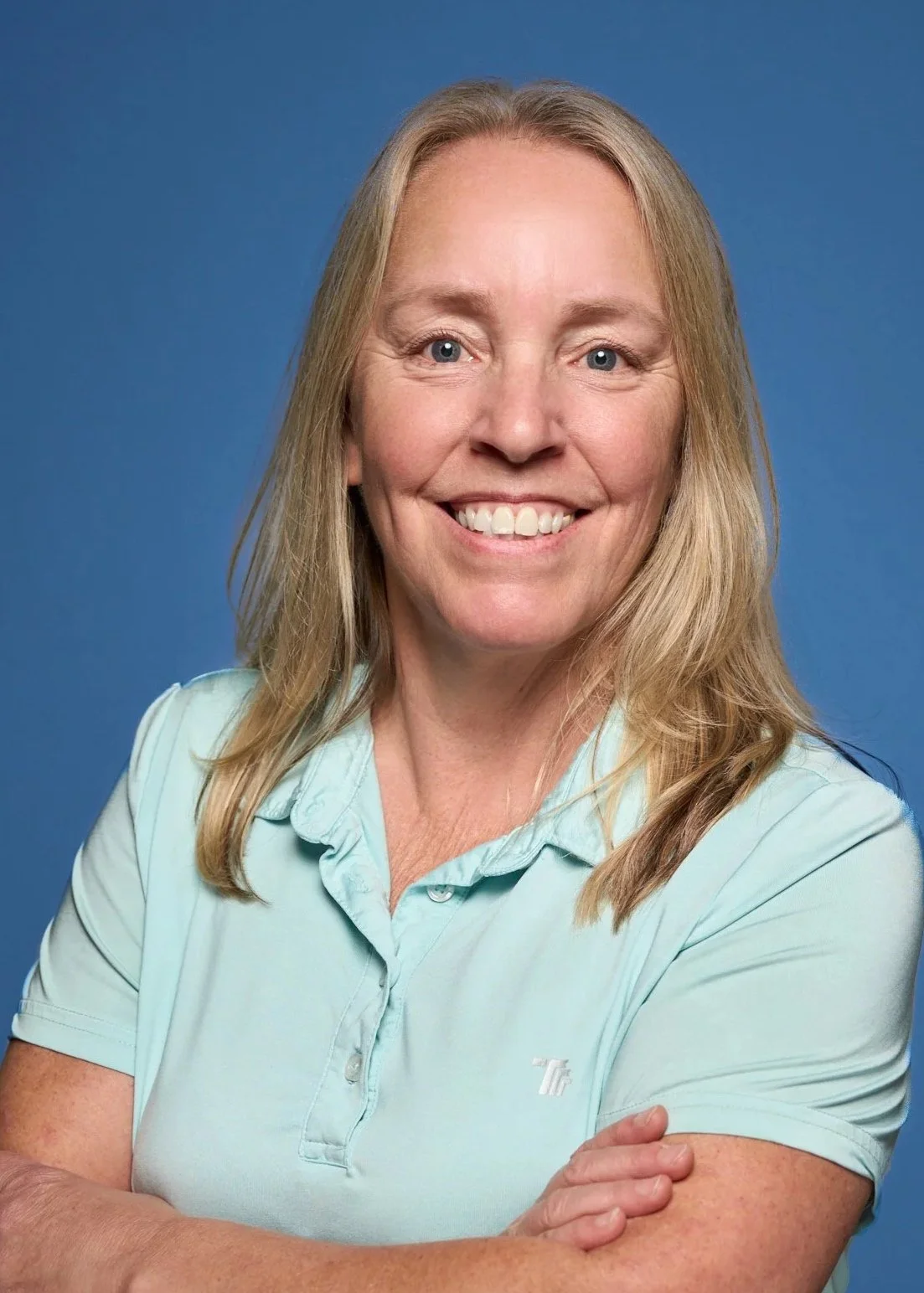 Portrait of a middle-aged woman with blonde hair, smiling, wearing a light blue polo shirt, against a blue background.