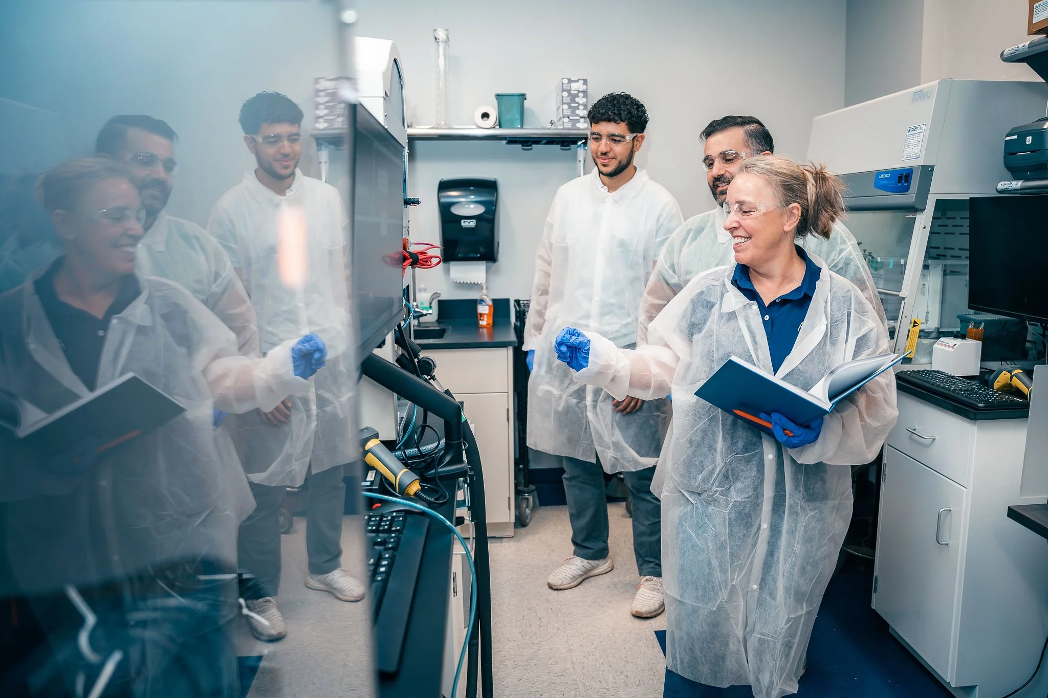 Group of scientists in lab coats and safety goggles working together in a laboratory, some holding notebooks and observing a computer screen.