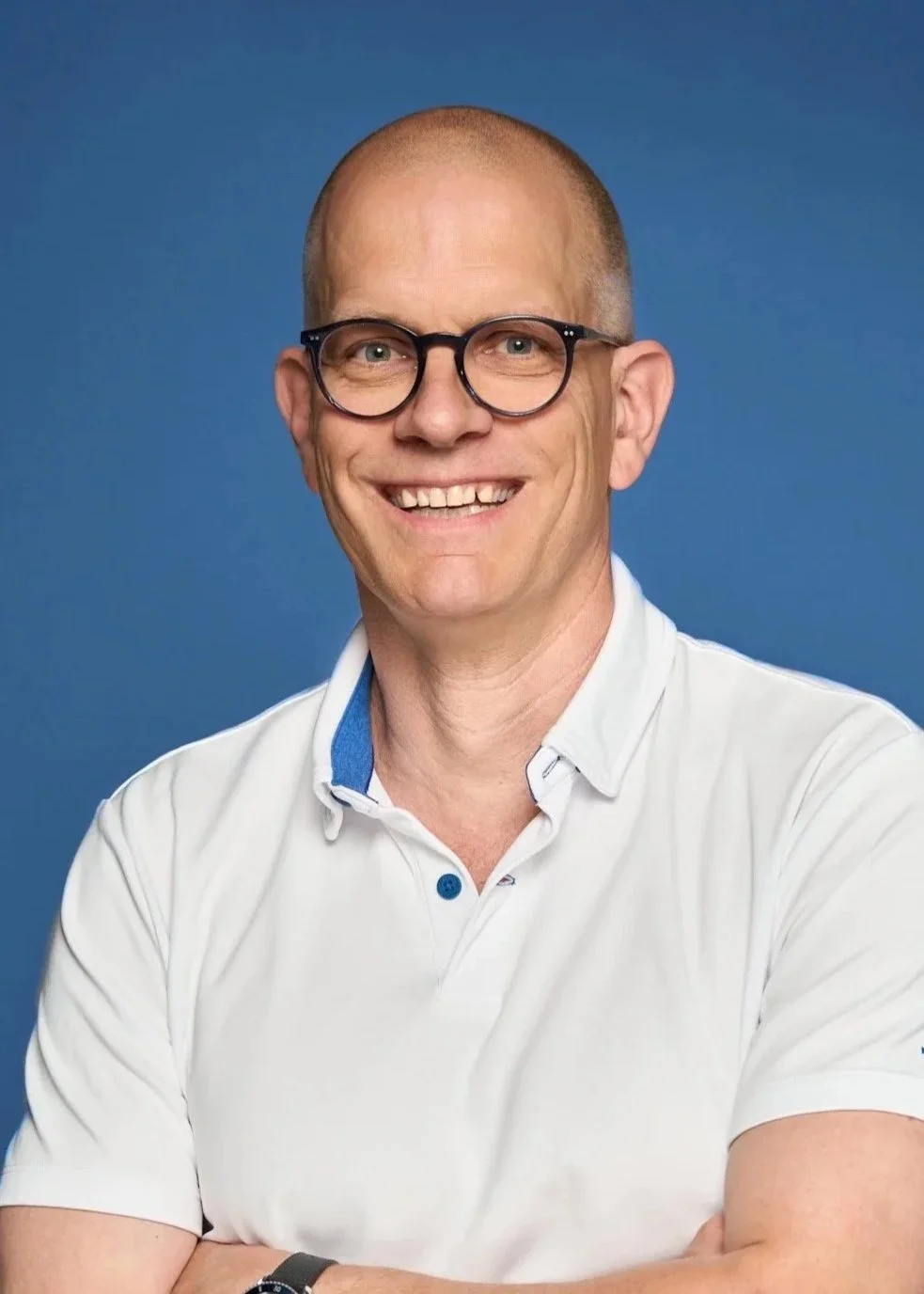 A smiling man with glasses and a white collared shirt standing against a blue background.