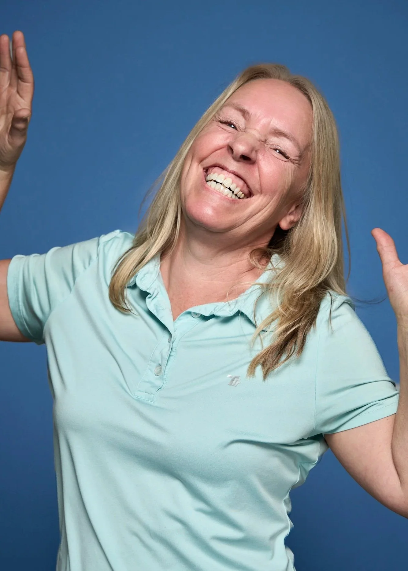 A woman with blonde hair smiling widely and making a playful gesture with her hands, wearing a light blue polo shirt against a blue background.