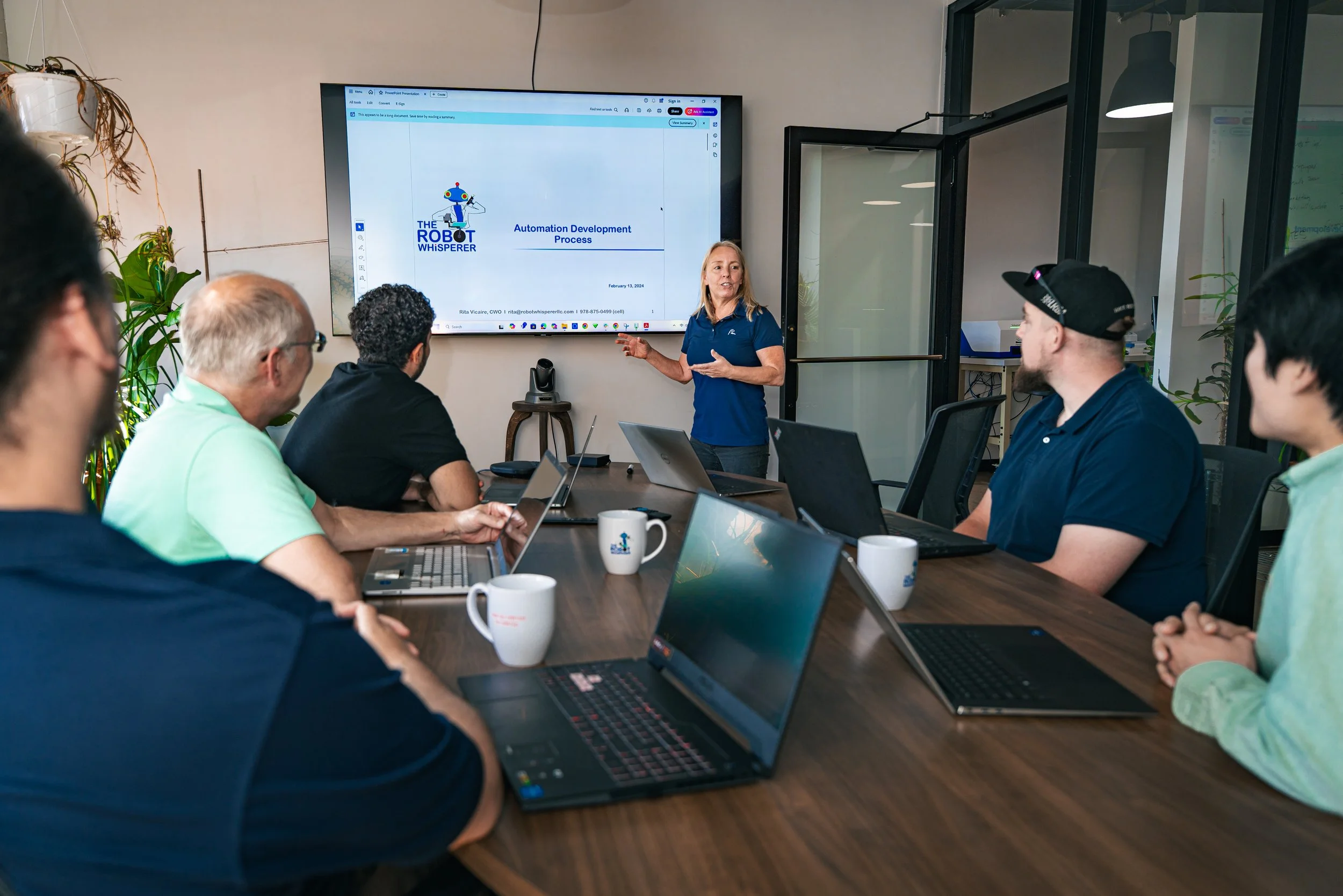 A woman giving a presentation to a group of six people seated at a conference table in a modern office. The presentation slide on the screen reads 'Automation Development Process'. The group is engaged, with laptops and coffee mugs in front of them.
