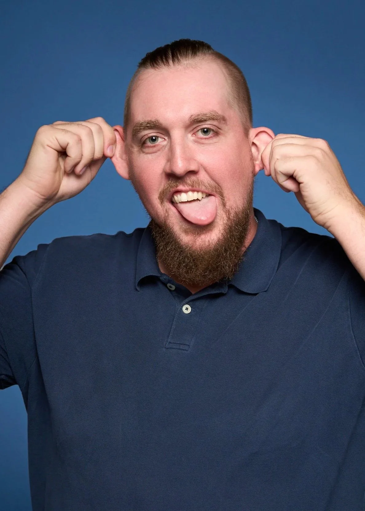 A man with light skin, a beard, and short hair, playfully sticking out his tongue and holding his hands up to his ears, wearing a navy blue polo shirt against a solid blue background.