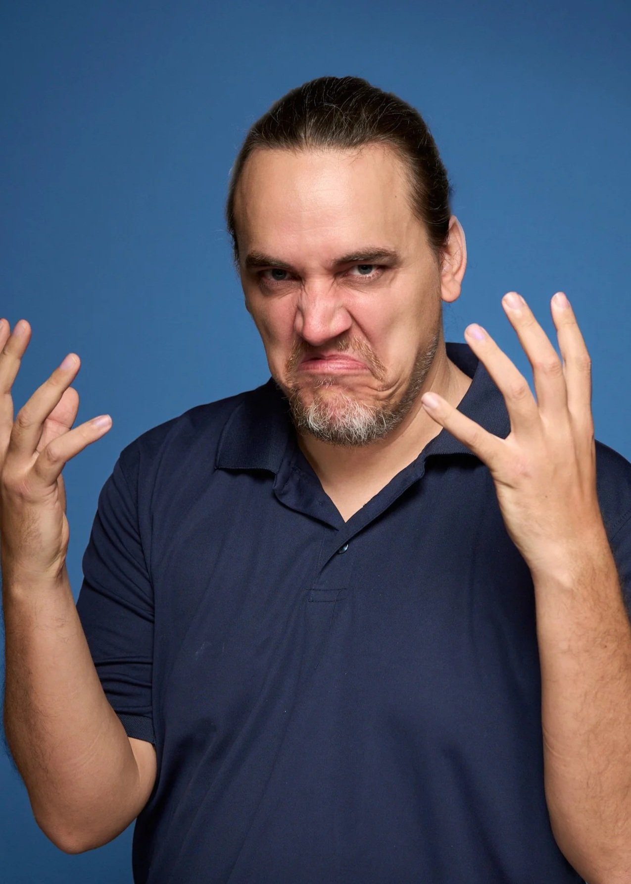 A man with long hair and a beard making a frustrated or angry facial expression, holding both hands up with fingers spread apart, against a blue background.