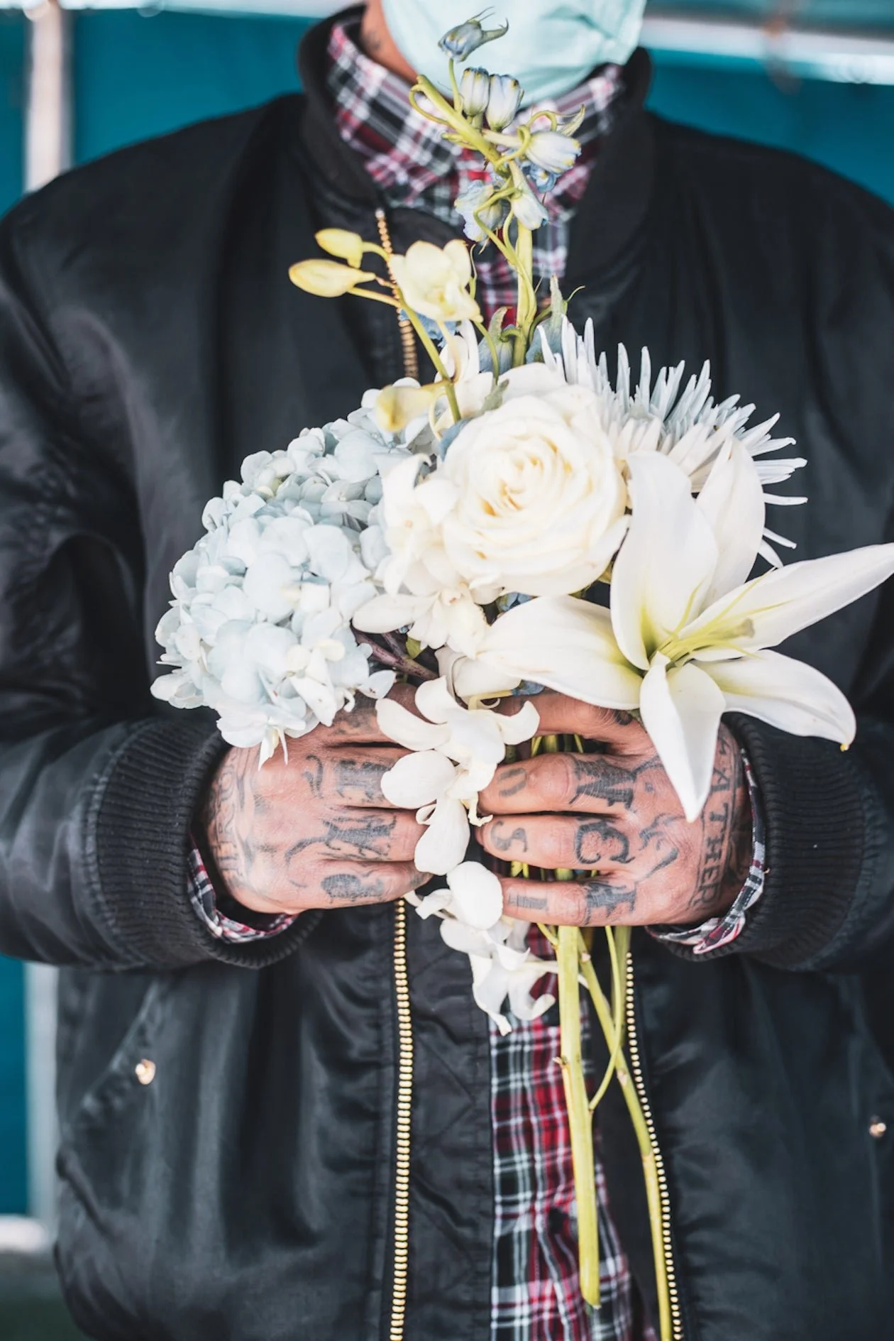 Person holding a bouquet of white flowers, including lilies and roses, wearing a black leather jacket, colorful plaid shirt, and a face mask.