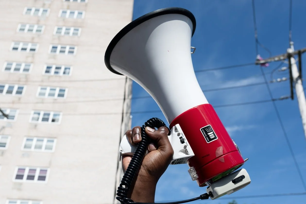 A person's hand holding a red and white megaphone pointed upward, with a city building and power lines in the background.