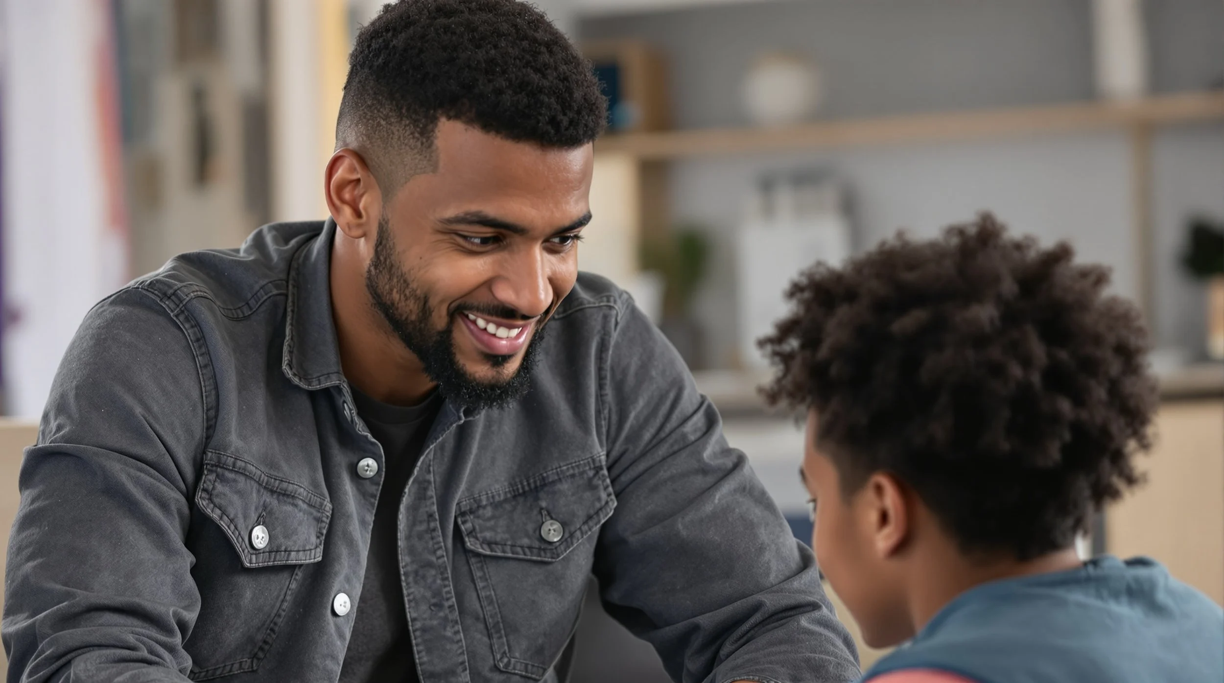 A man with dark hair and a beard, wearing a grey denim jacket, smiling and talking to a boy with curly hair.
