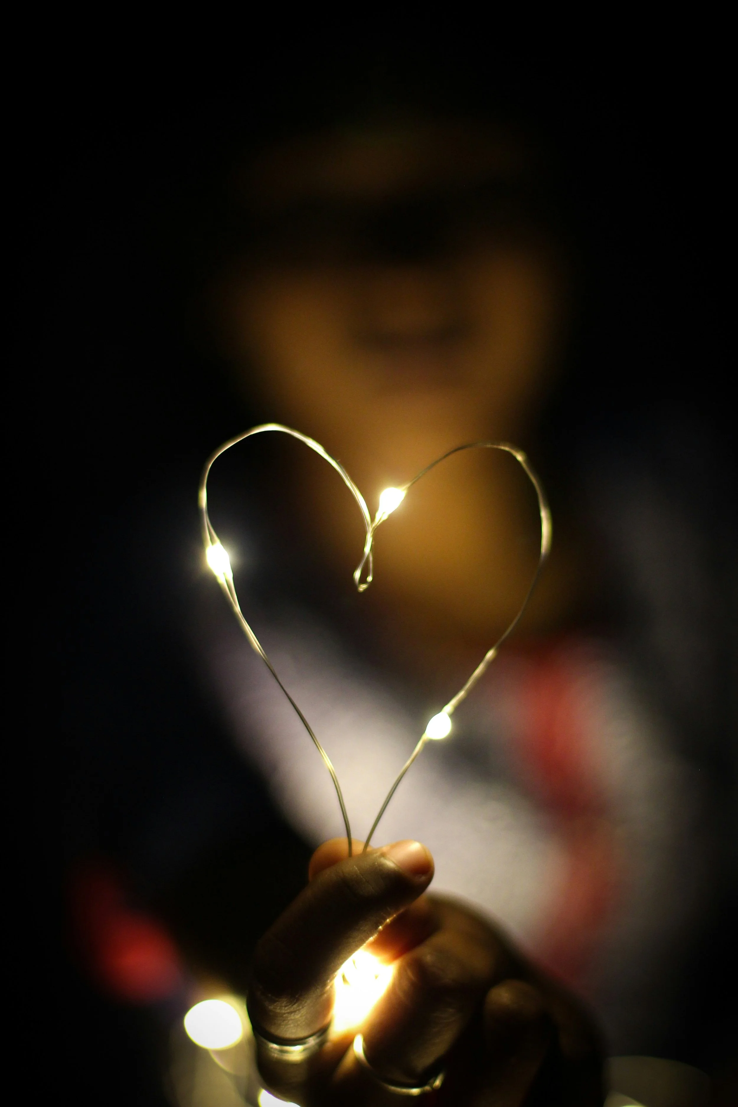 Person holding a string of fairy lights shaped like a heart with blurred face in background.