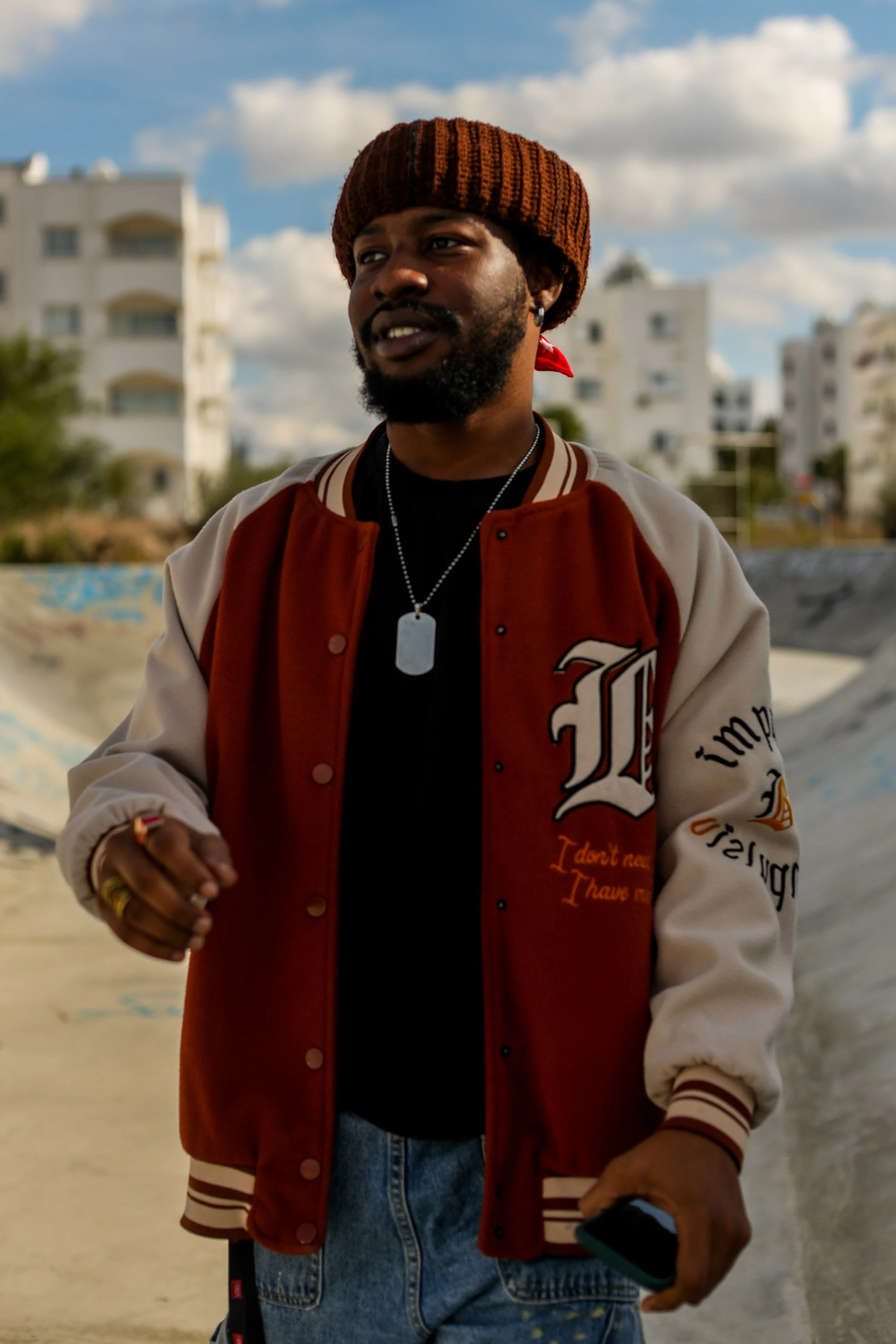 A man wearing a brown knit beanie, a brand baseball jacket, a black shirt, jeans, and jewelry standing outdoors with buildings and cloudy sky in the background.