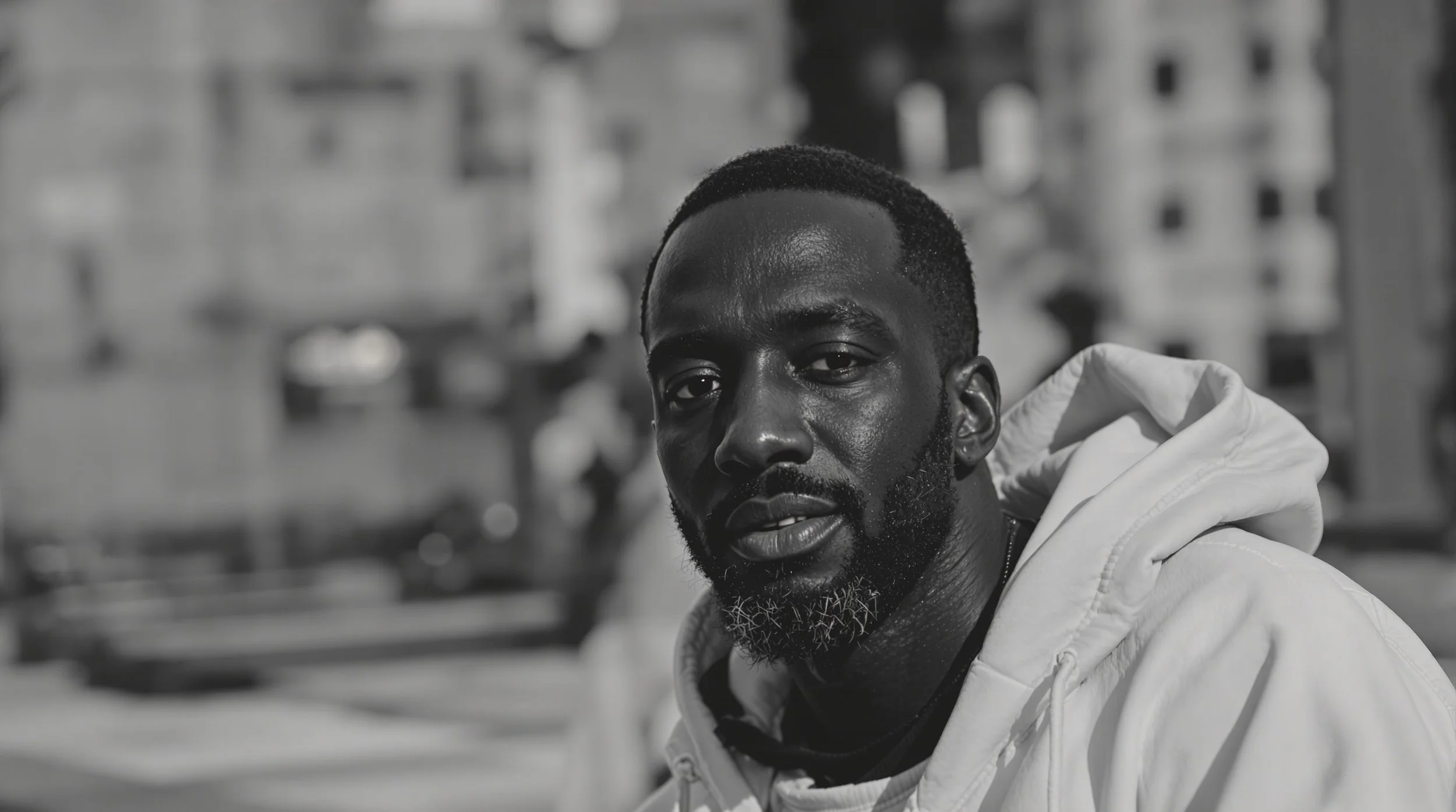 A young Black man with a short beard and short haircut, wearing a white hoodie, looking directly at the camera with a cityscape background in black and white.