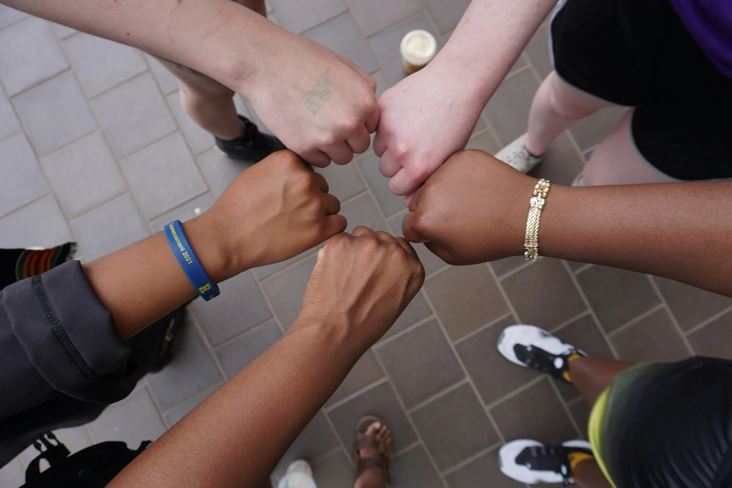 Six people standing in a circle making fists together in the center, viewed from above on a tiled floor.