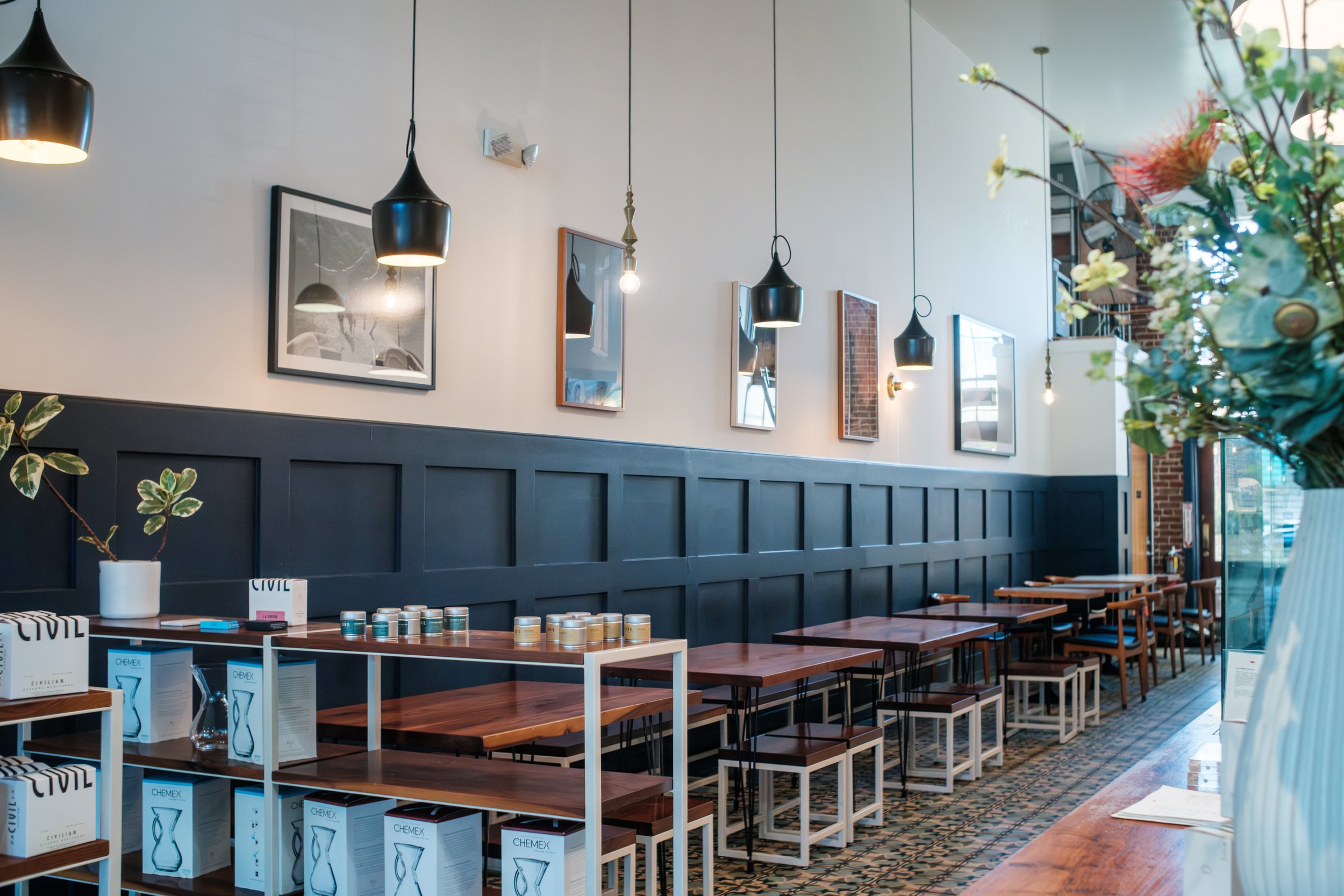 Interior of a modern cafe or restaurant with black and wood decor, multiple black pendant lights, framed art on the wall, tables with chairs, plants, and display shelves with products, including Chemex coffee makers.