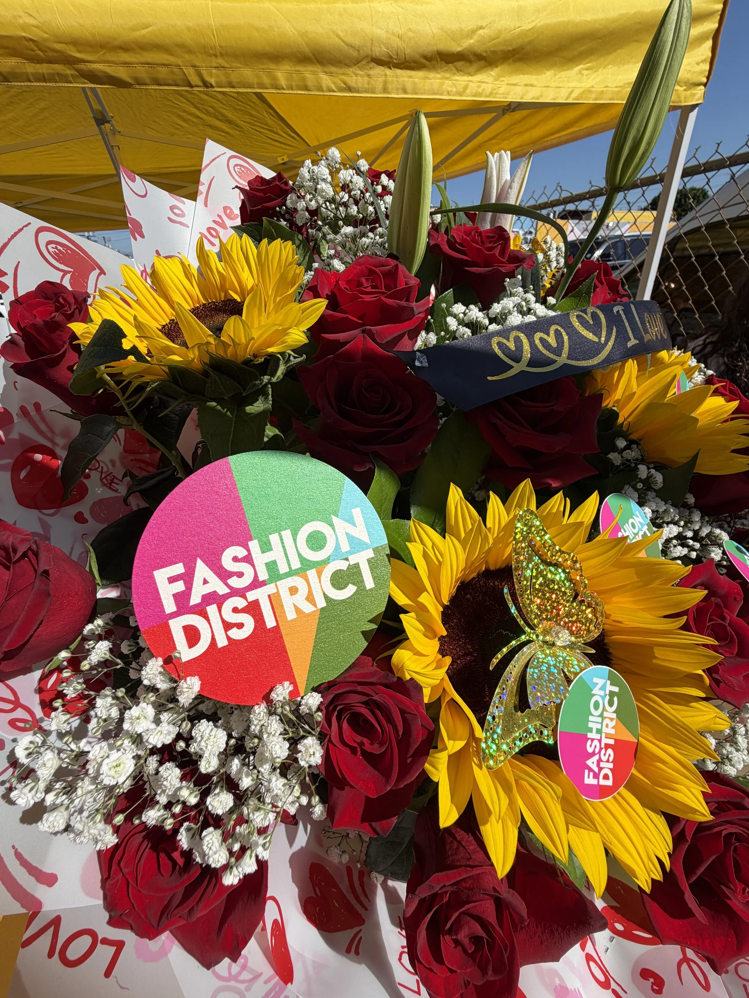 A colorful bouquet of red roses, sunflowers, white lilies, and baby's breath with heart-shaped "Love" signs and a black ribbon that says "Love" in gold. There is a yellow canopy overhead and a chain-link fence in the background.