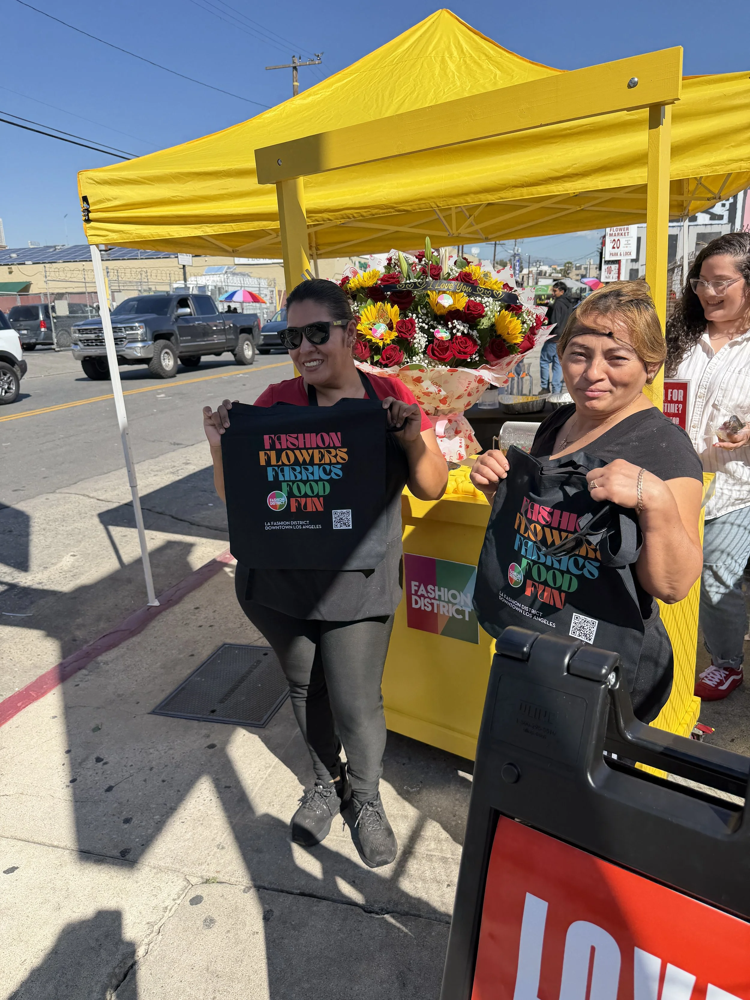 Woman holding a black shopping bag with colorful text standing in front of a yellow vendor stall at an outdoor market on a sunny day.