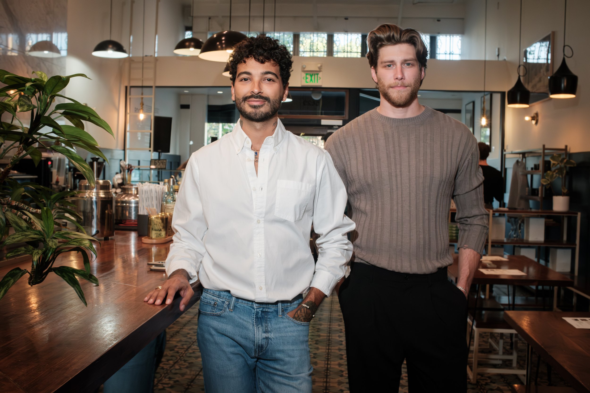 Two men standing side by side in a well-lit modern interior, with one resting a hand on a wooden counter, both looking at the camera. The man on the left is wearing a white shirt and has curly dark hair, while the man on the right is wearing a gray sweater and has wavy brown hair.