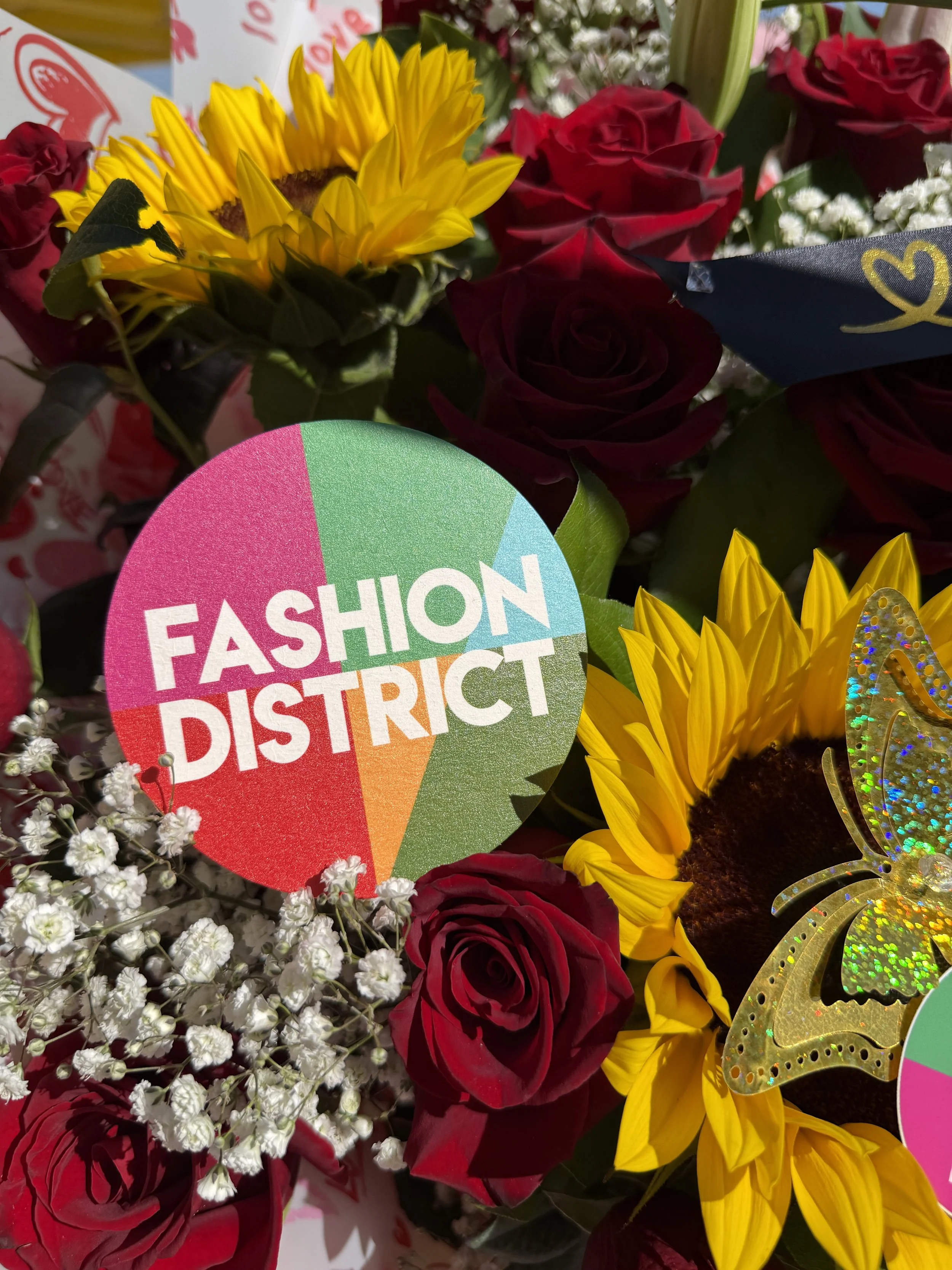 A colorful bouquet of flowers including red roses, yellow sunflowers, white baby's breath, and small white flowers, with a sticker that reads 'FASHION DISTRICT'.