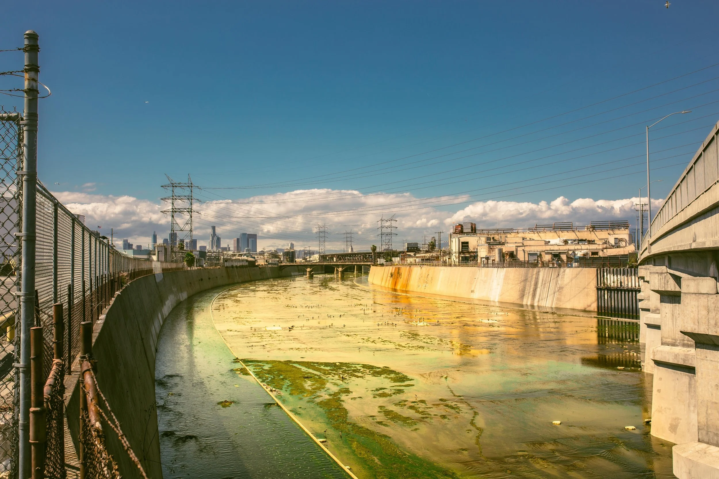 Cityscape with a flood control channel filled with green algae, a fence on the left, buildings in the distance, power lines, and a partly cloudy sky.