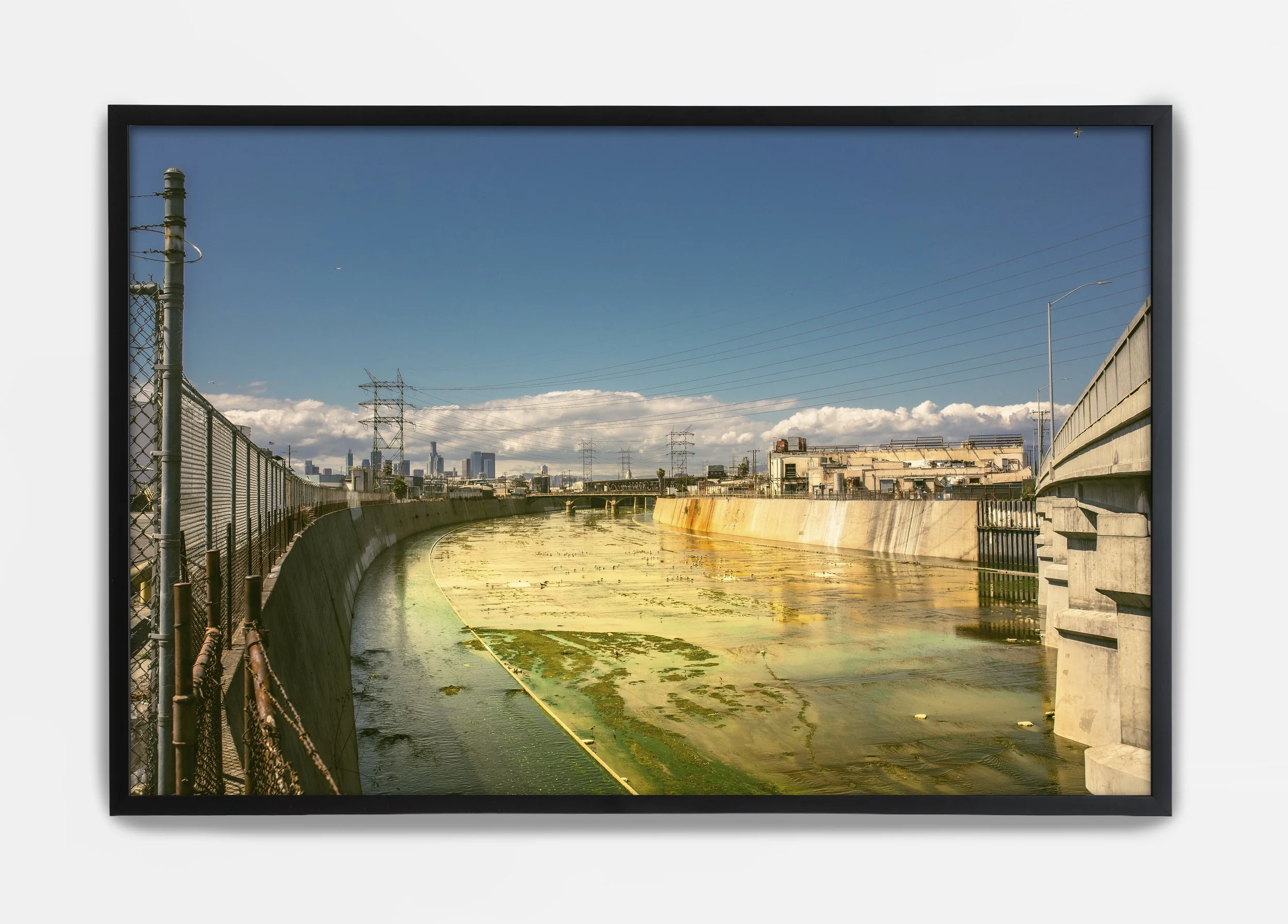 A framed photograph of an urban canal with a city skyline in the background, power lines, and a chain-link fence along one side, under a blue sky with some clouds.