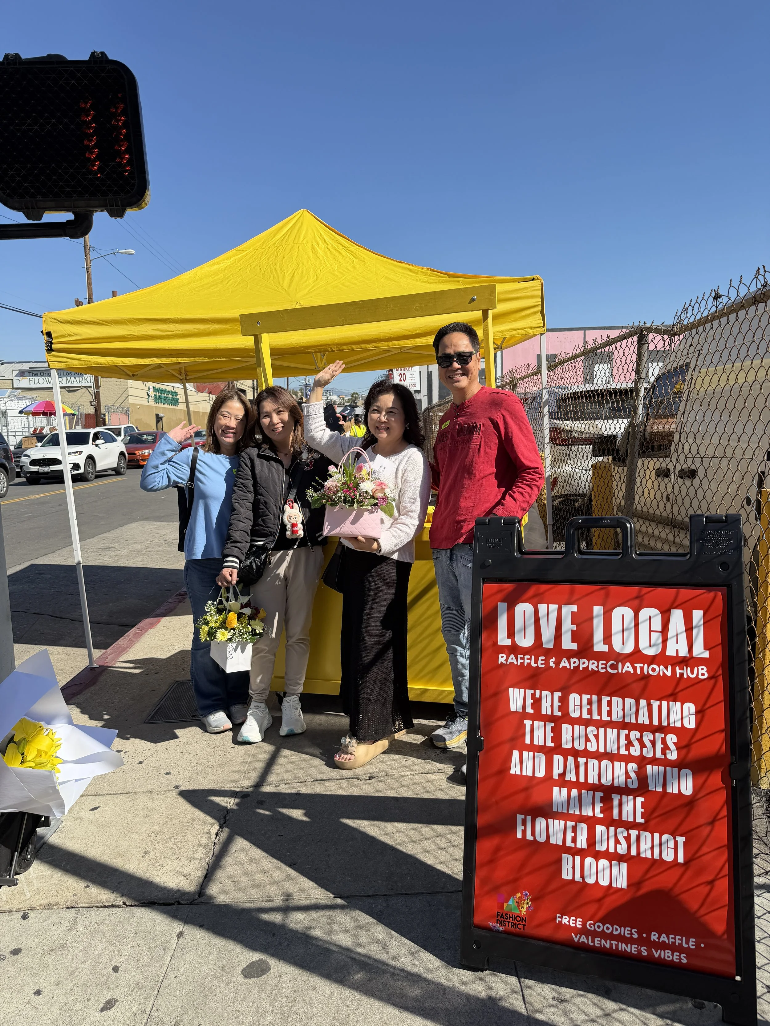 Four people smiling and posing in front of a yellow tent at an outdoor event, with a sign that reads 'Love Local Raffle & Appreciation Hub' and mentions celebrating local businesses and patrons, with parked cars and buildings in the background.