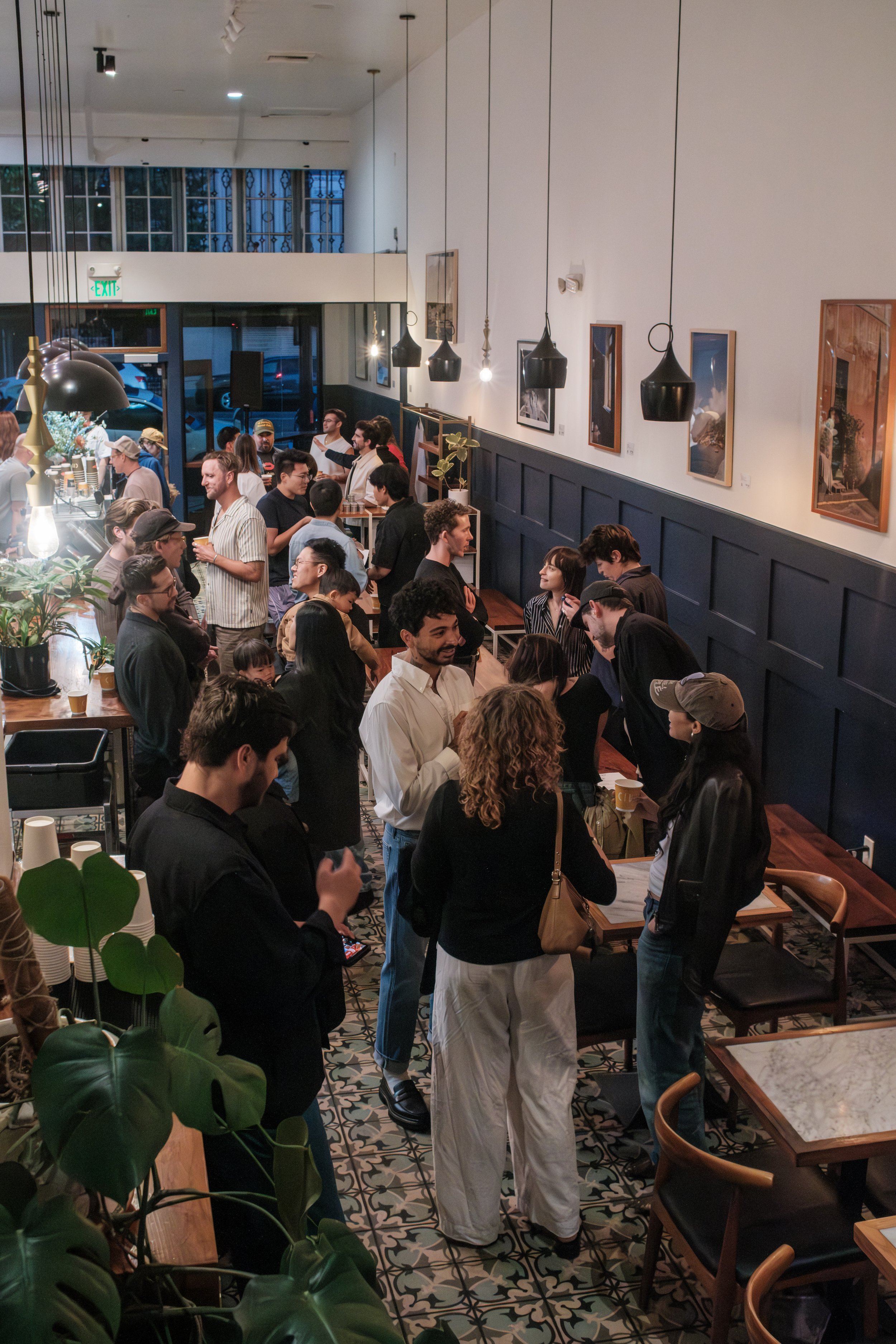 People gathering and standing in a busy cafe or restaurant with artwork on the walls and patterned tile flooring.