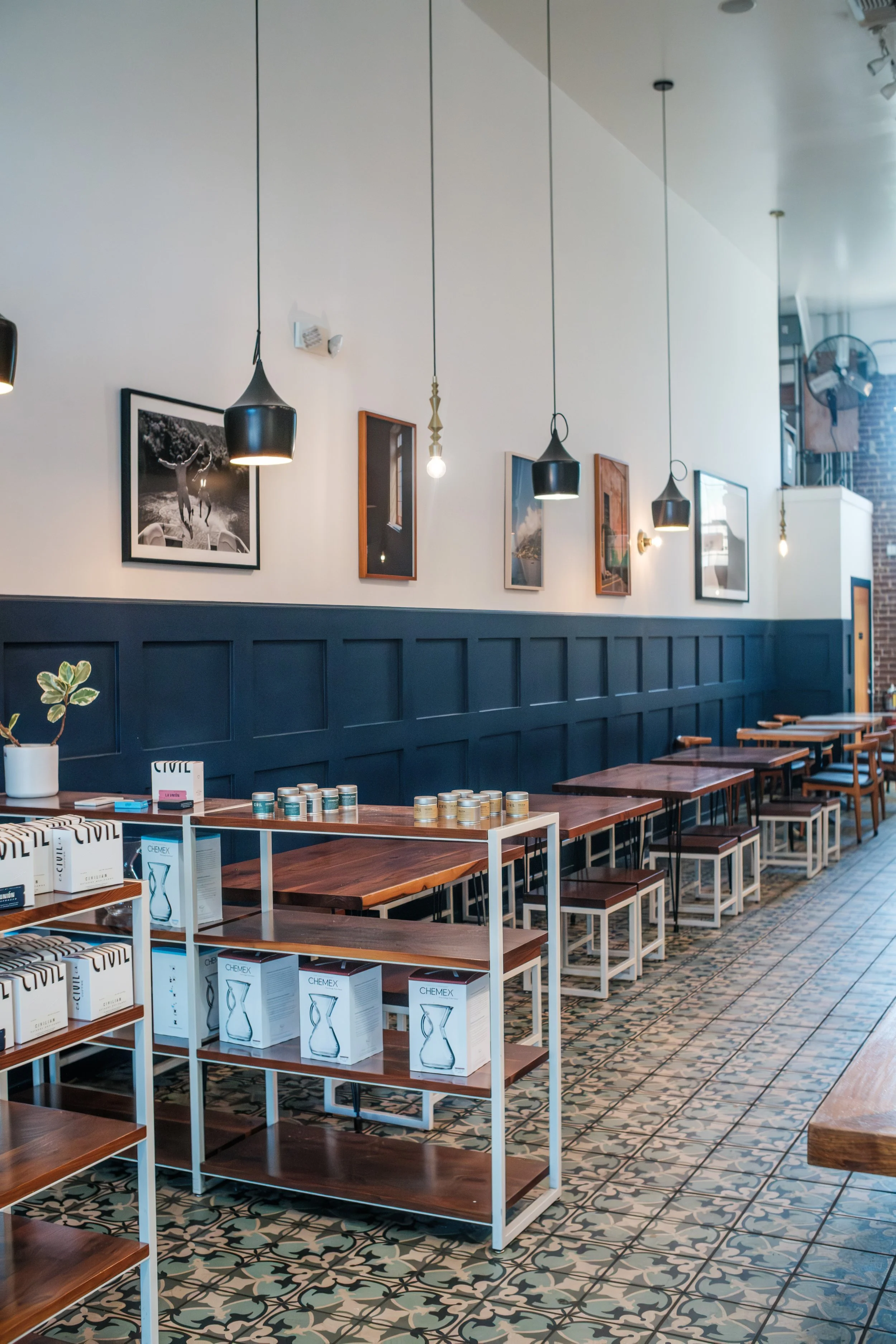 Interior of a modern cafe or restaurant with wooden tables and white shelving displaying boxed glassware and jars. The upper wall has a dark blue wainscoting, and the walls are decorated with framed photographs. Pendant and bare bulb lighting hang from the ceiling, and there is patterned tile flooring.