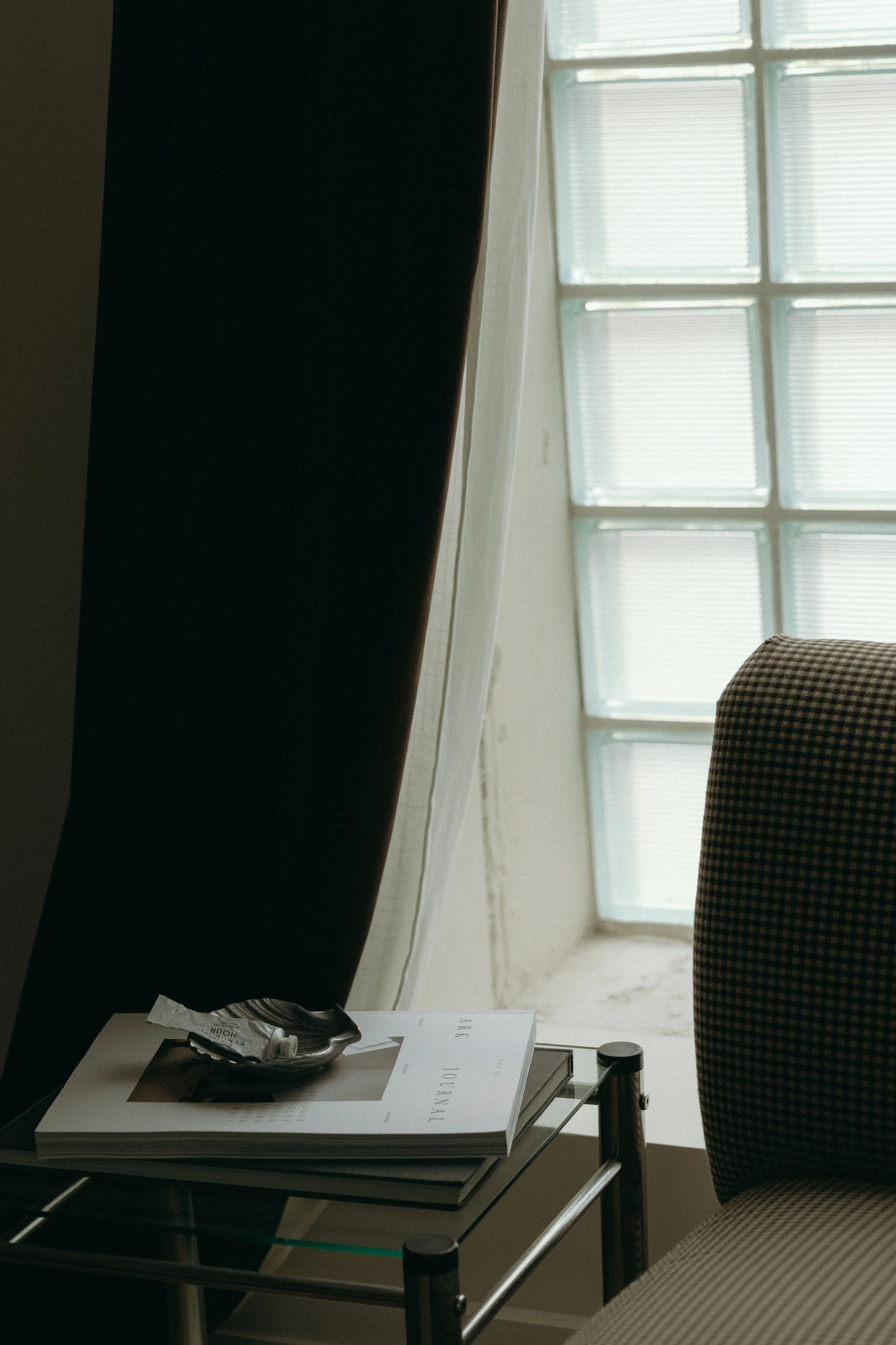 A glass table with a magazine and a small black dish holding a crumpled wrapper, next to a beige and black checkered armchair, with a window made of glass blocks in the background.