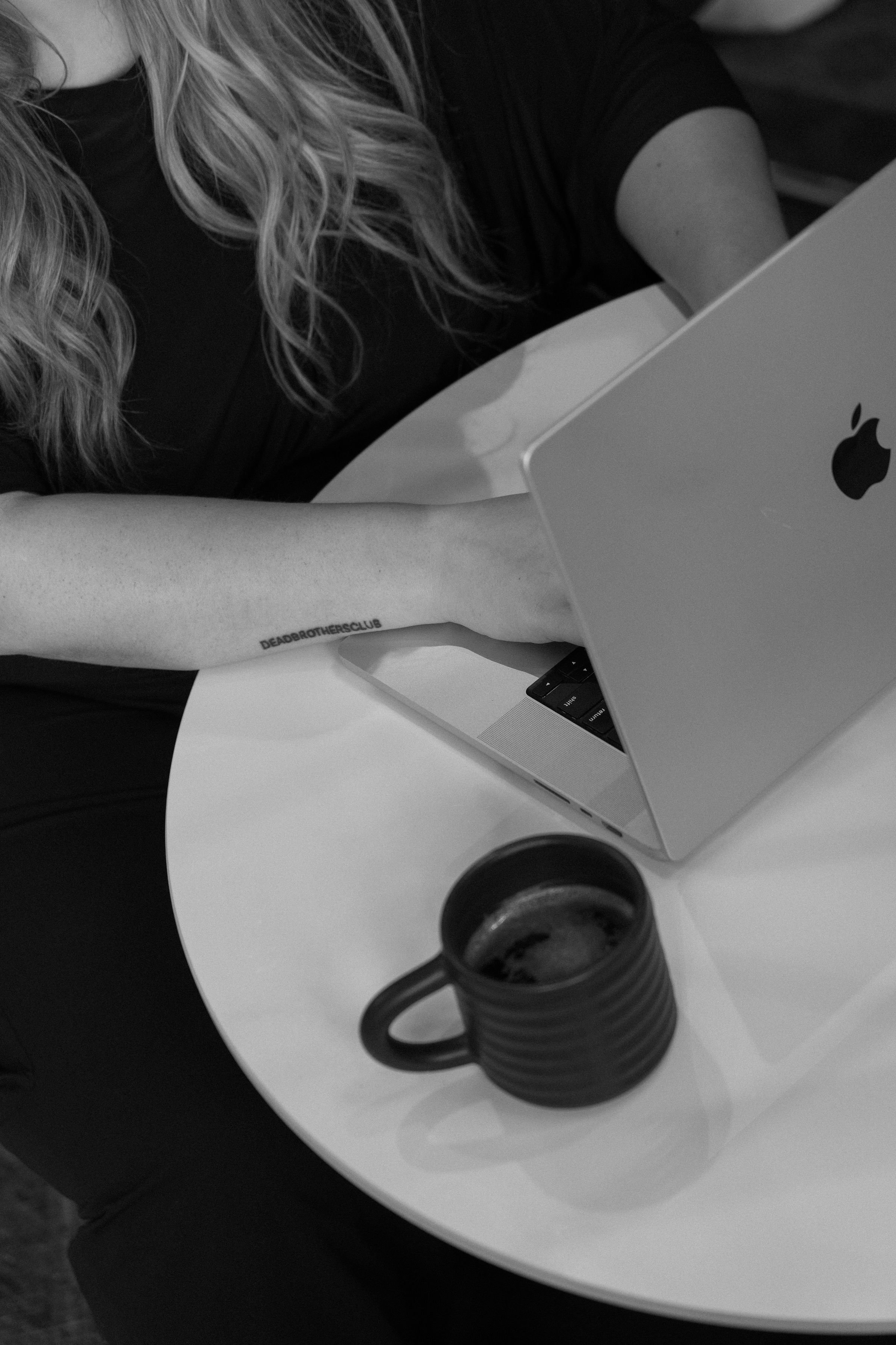 Person with long wavy hair working on a MacBook with a coffee mug on a white table.