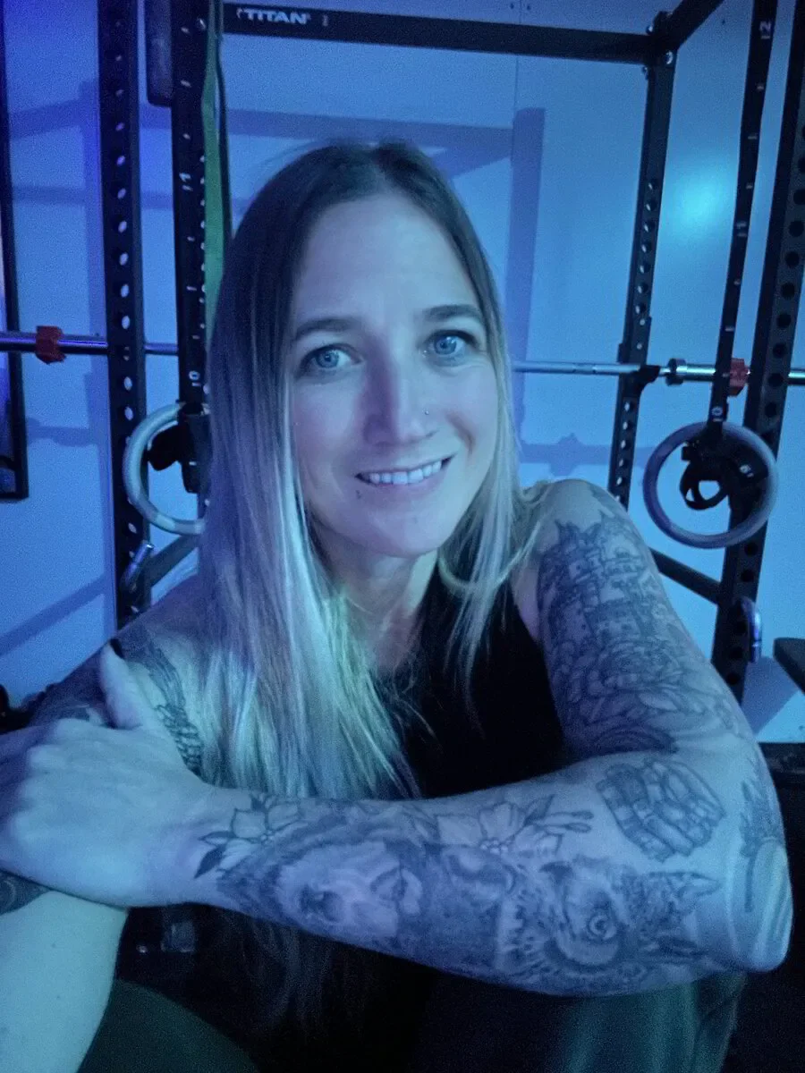 A woman with long blonde hair and tattoos on her arms taking a selfie in a gym, with weightlifting equipment and gym rings in the background.