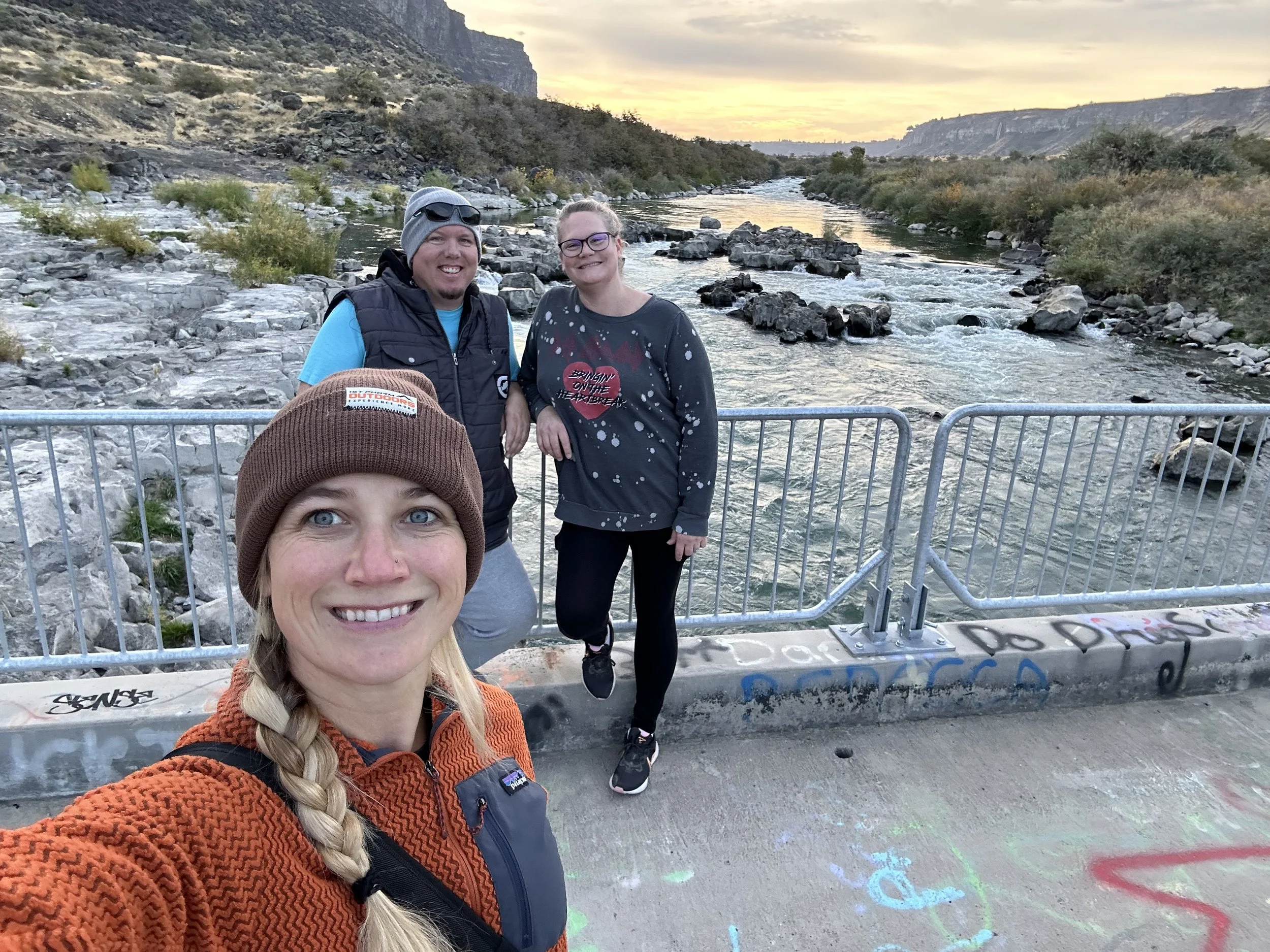 Three smiling friends taking a selfie on a bridge over a river at sunset. The person in front has blonde hair in a braid, wearing a brown hat and orange jacket. The person on the left is wearing glasses, a gray beanie, and a black vest. The person on the right has glasses, blonde hair, and is wearing a dark sweatshirt. Graffiti is visible on the bridge's concrete surface, with a scenic river and rocky landscape in the background.