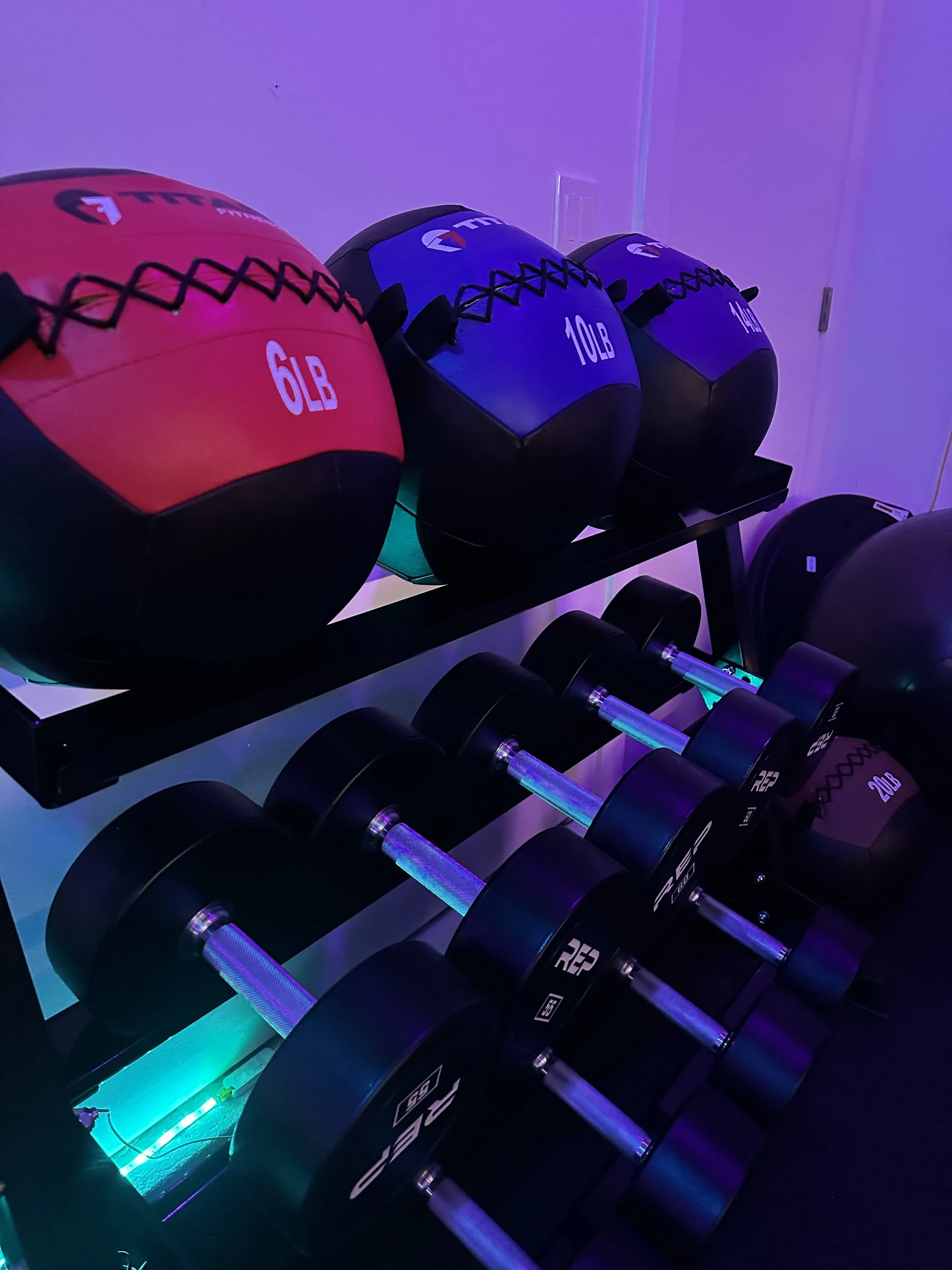 Set of medicine balls and dumbbells with glowing lights in a fitness room, including a red 6 lb ball, a green 10 lb ball, and a purple 15 lb ball, on black racks under purple lighting.