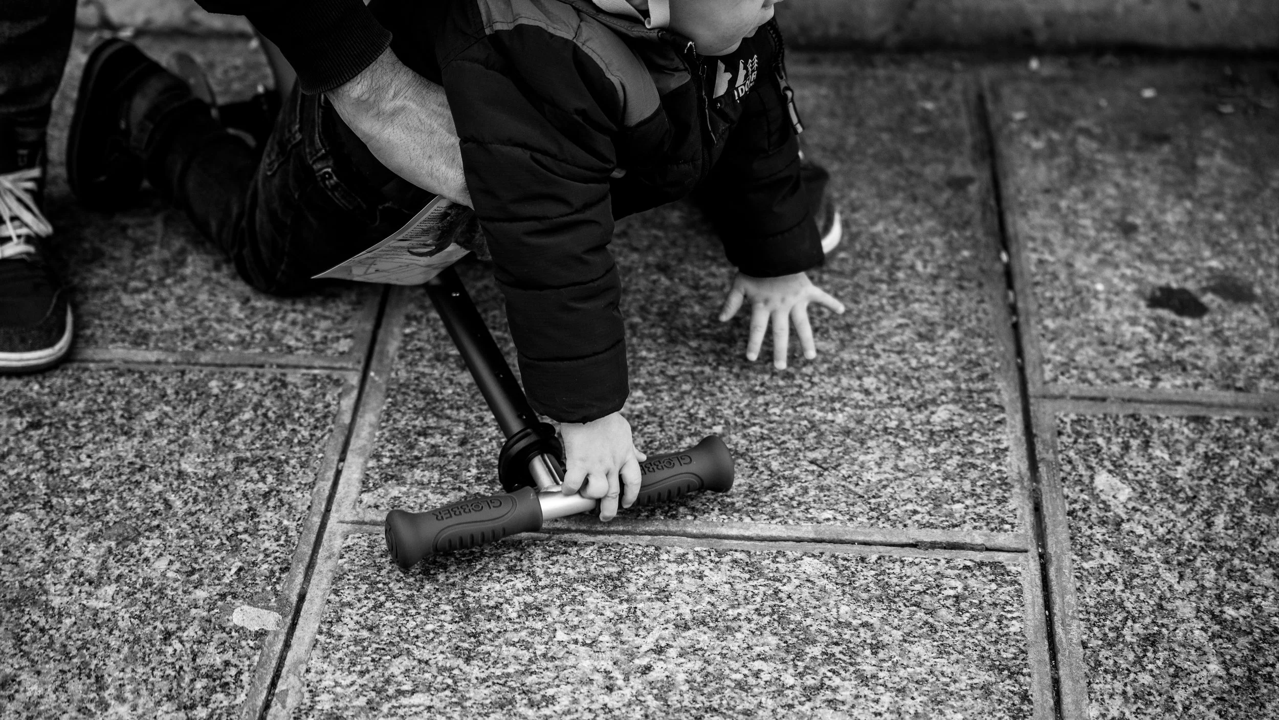 A young child on the ground, on their hands and knees, holding a scooter, wearing a dark jacket and shoes, next to a paved sidewalk.