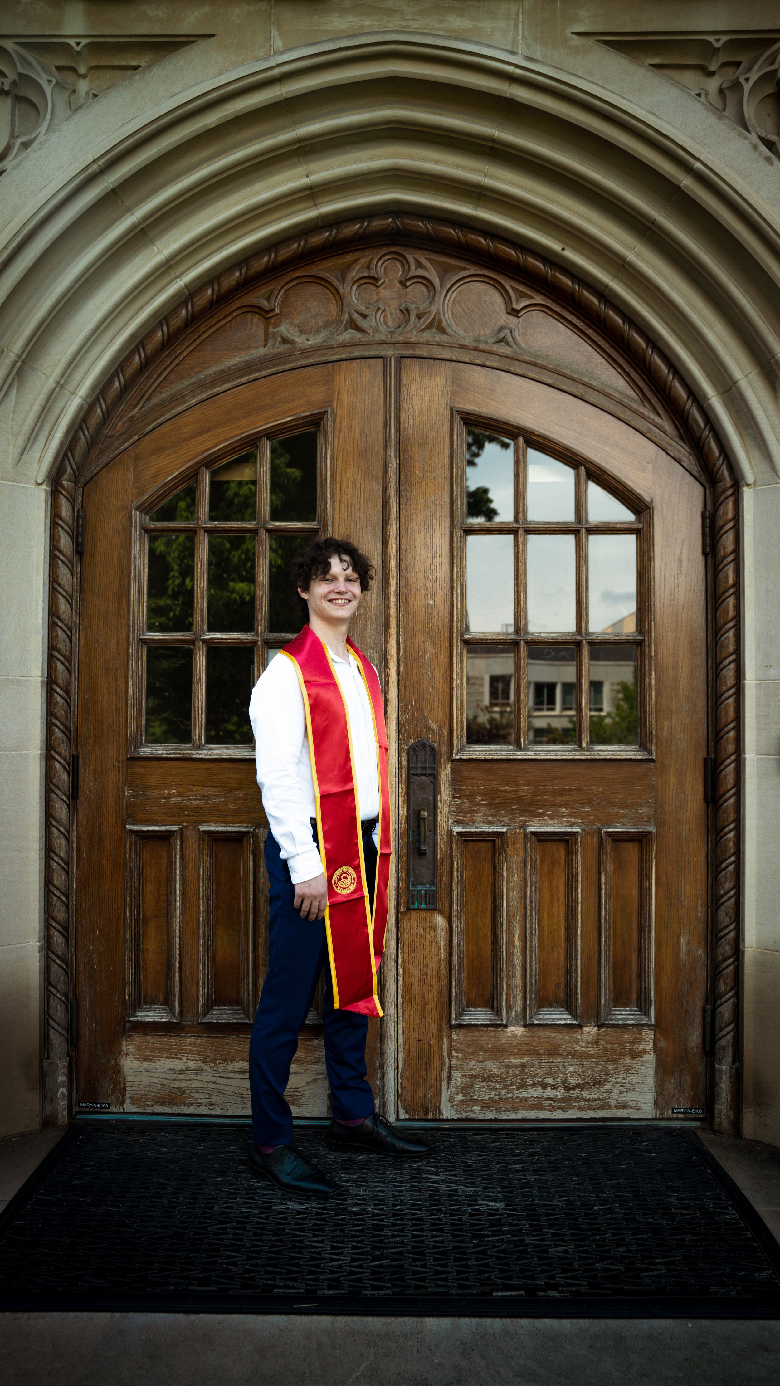 A young man in formal attire with a red stole standing in front of a large wooden door.