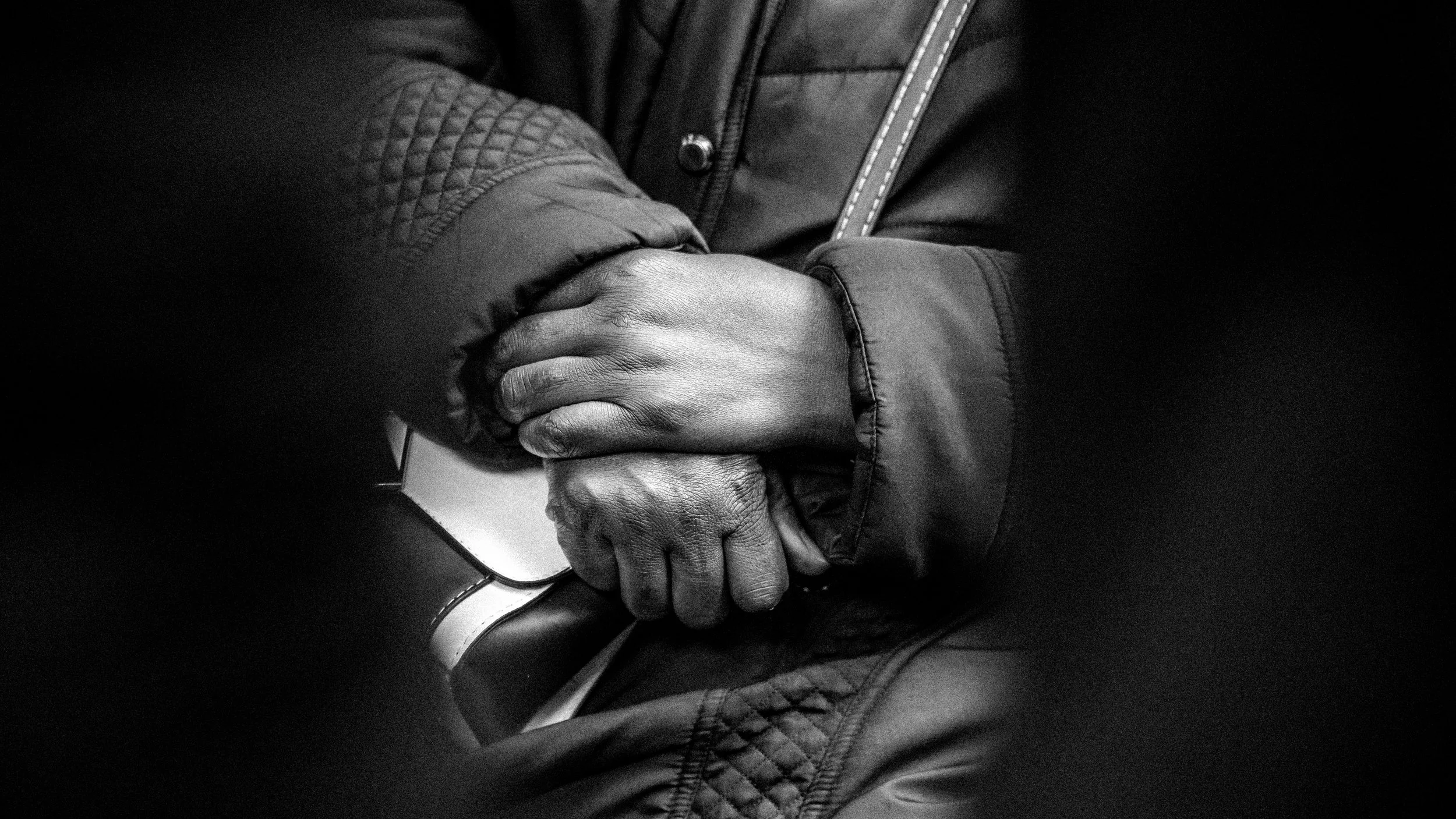 Close-up of two hands clasped together, resting on someone's lap, with the person wearing a jacket with quilted sleeves.