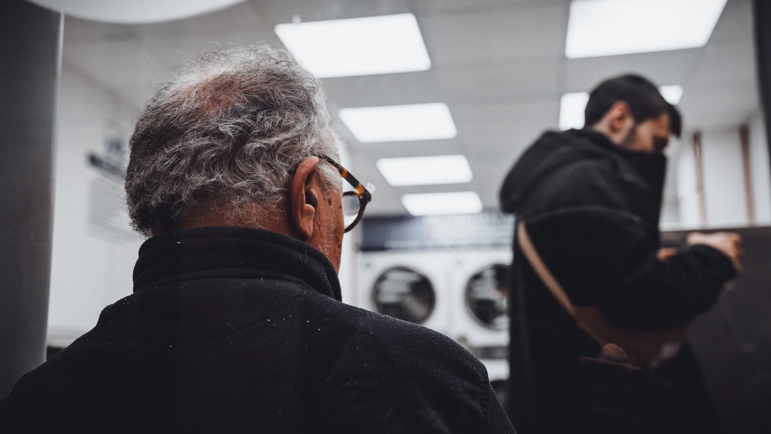 Older man with gray hair and glasses sitting in a laundromat, with a younger man in a black hoodie using a machine nearby.