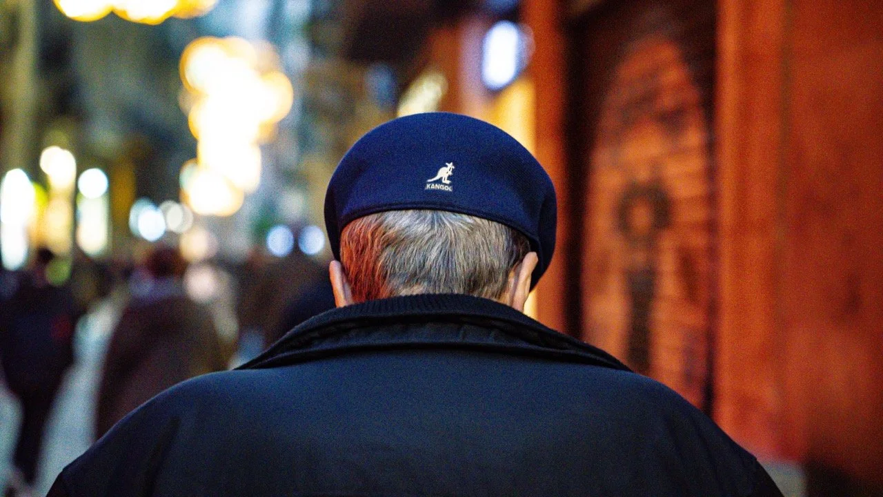 Back view of a person wearing a navy blue beret and black coat, walking in a city street at night with blurred lights and brick buildings in the background.