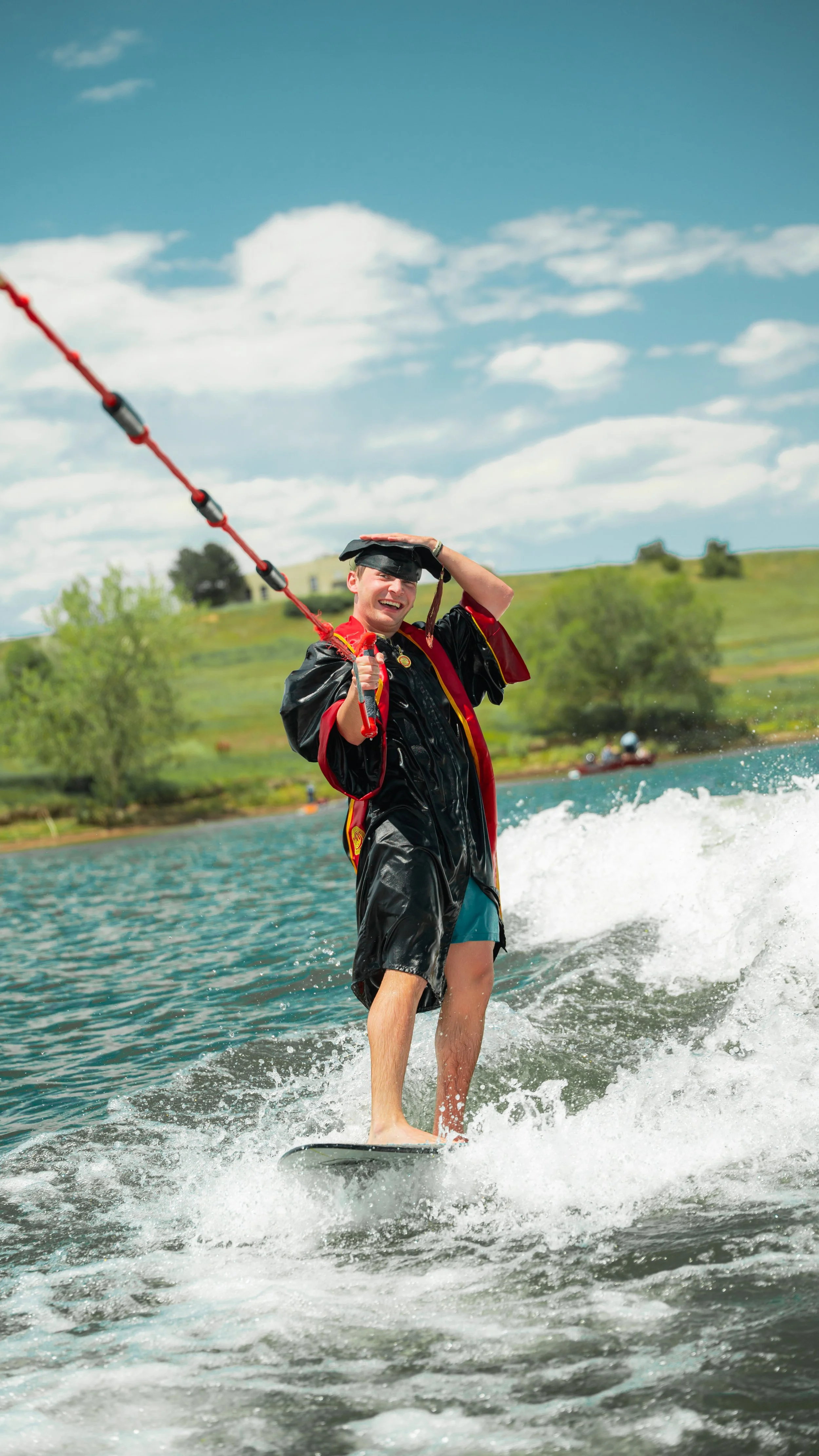 A young man in a graduation cap and gown, wearing a life jacket, wakesurfing on a lake under a partly cloudy sky with green hills in the background.