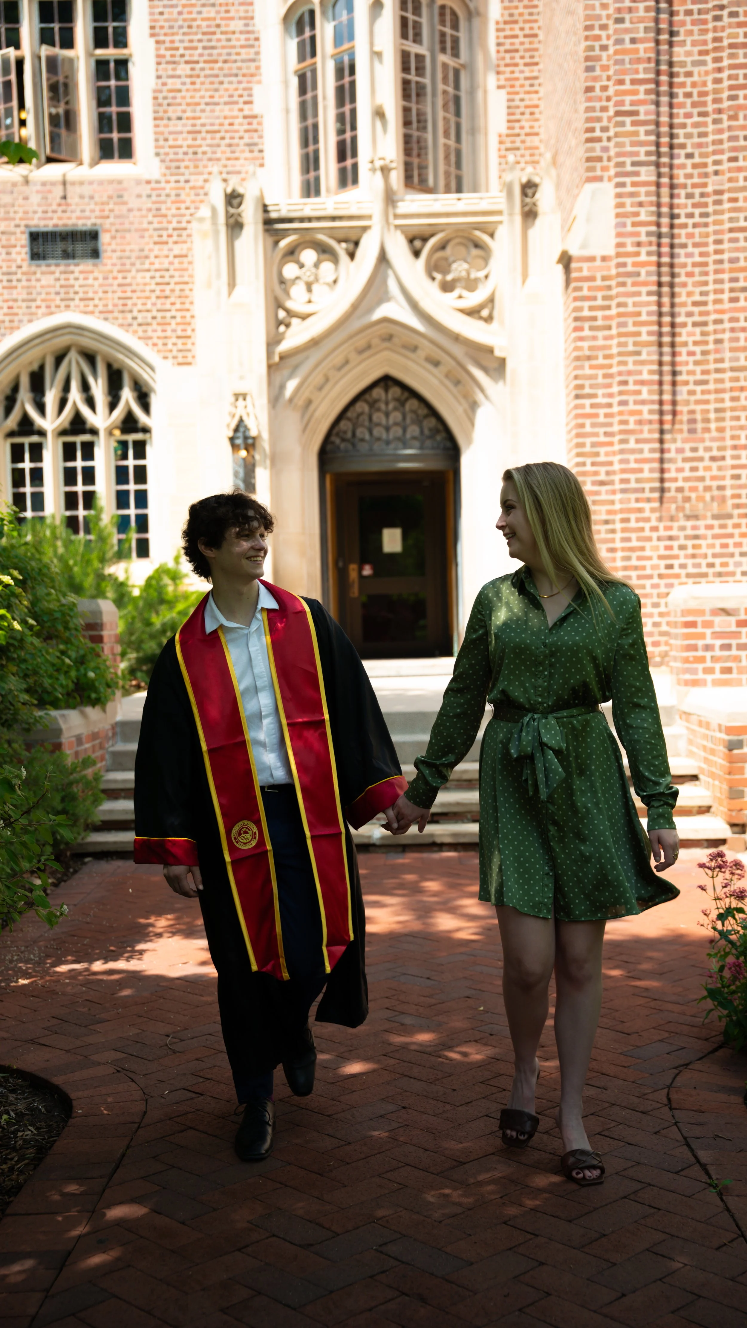 A graduate in a black cap and gown with red and yellow trim holding hands with a woman in a green dress, walking on brick pavement in front of a brick and stone building with large windows.
