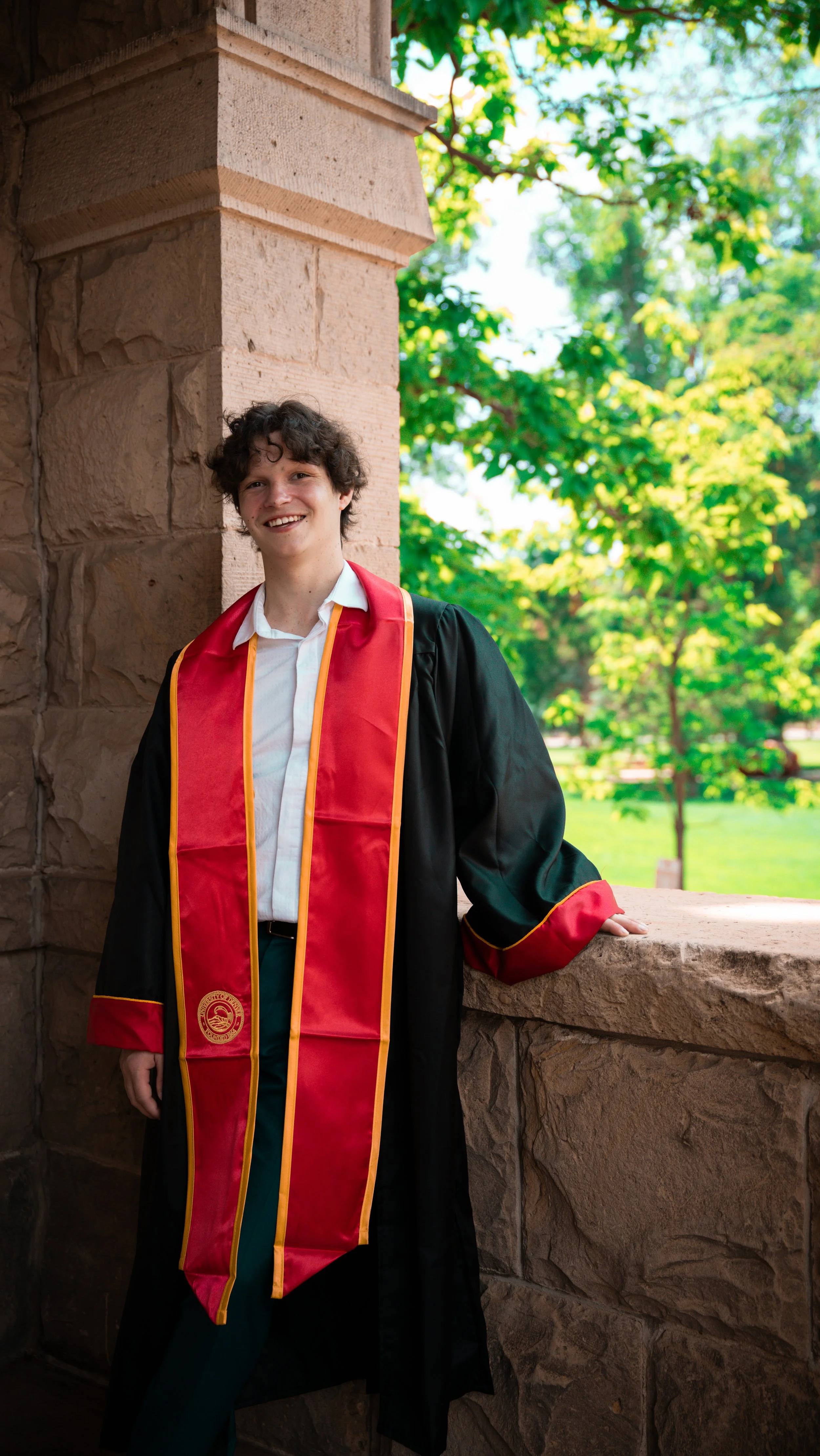 Young man in graduation gown and red stole, standing outdoors on a stone balcony with lush green trees in the background, smiling.