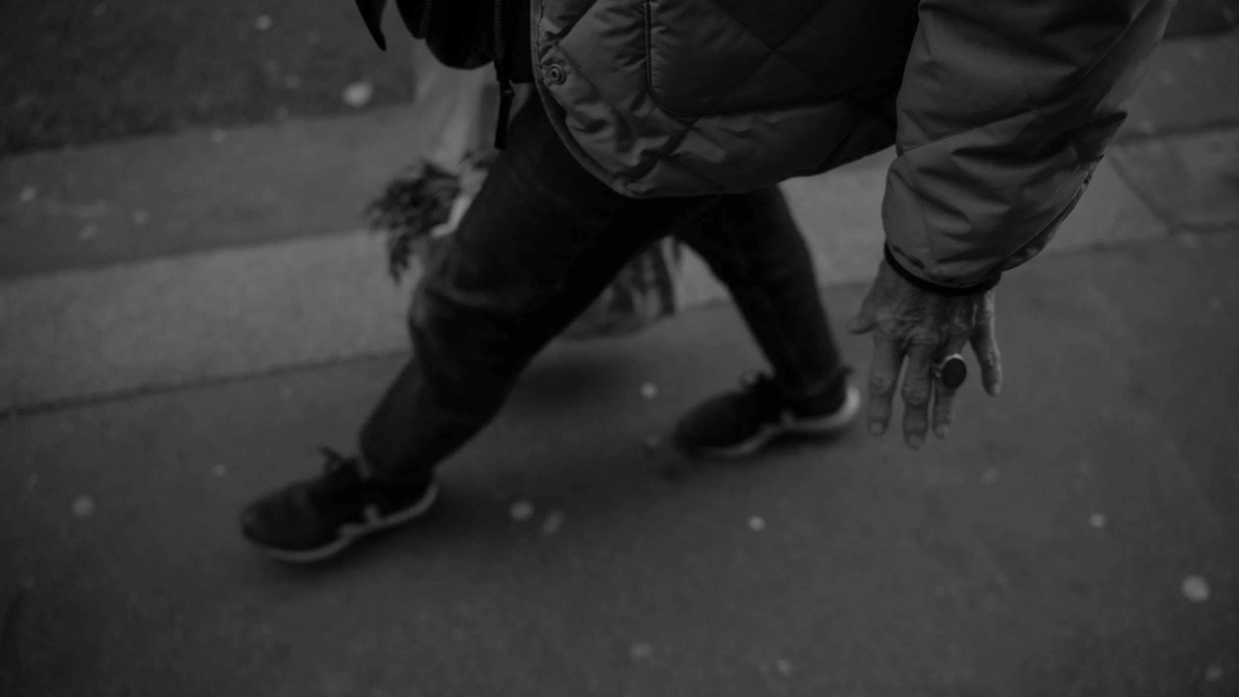 A person in a jacket crouching in a street, with hand extended toward the ground, on a sidewalk.