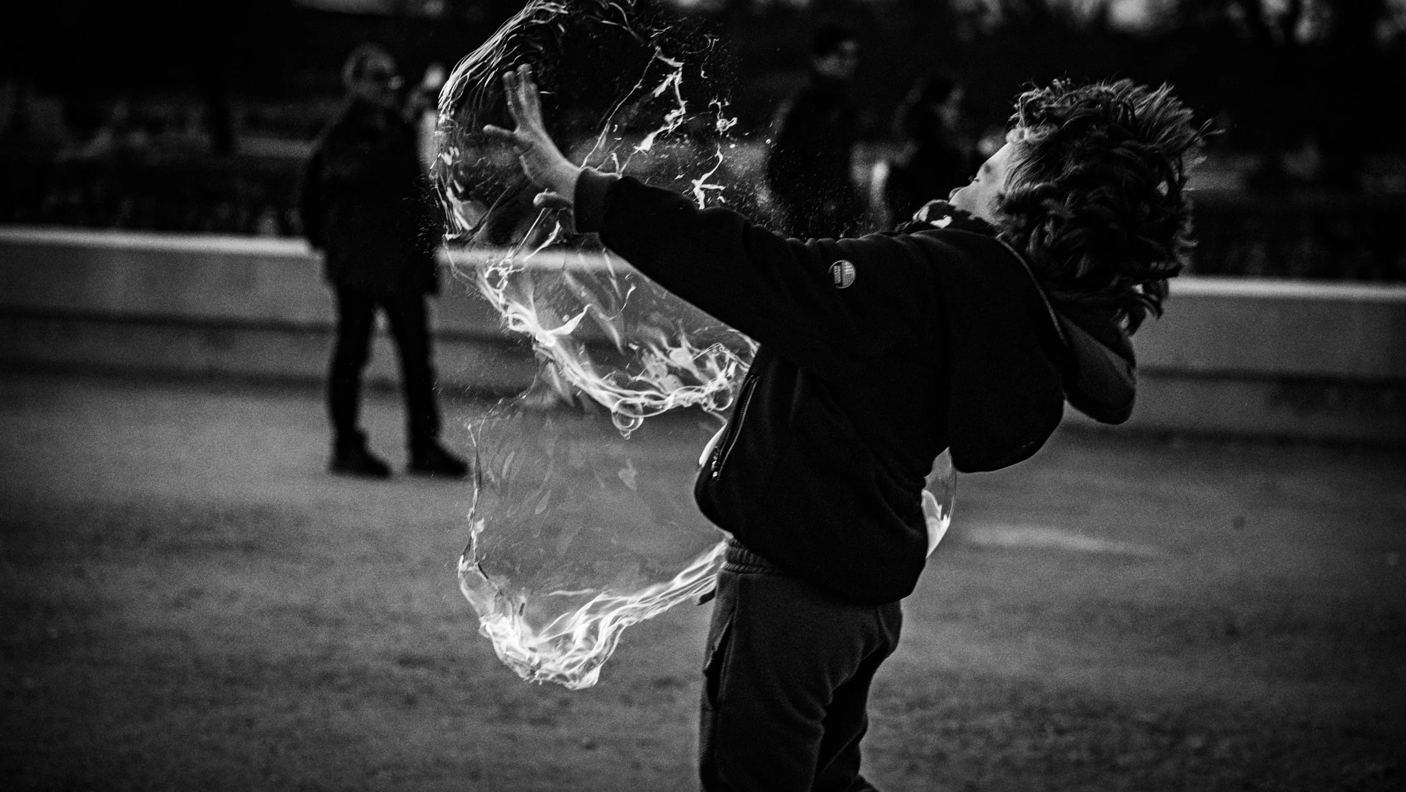 A person with curly hair is playing with a large soap bubble in an outdoor setting at night. There are two other people in the background, slightly out of focus.