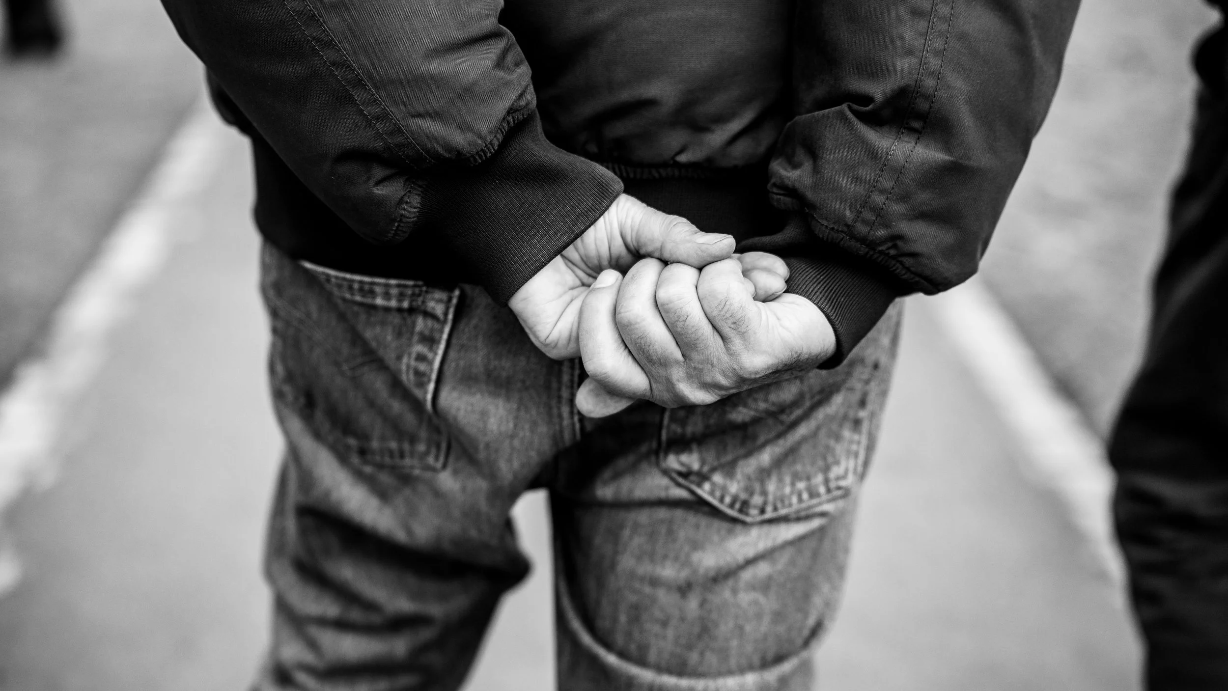 Black and white photo of a person with hands clasped behind their back, wearing a jacket and jeans, standing outdoors.