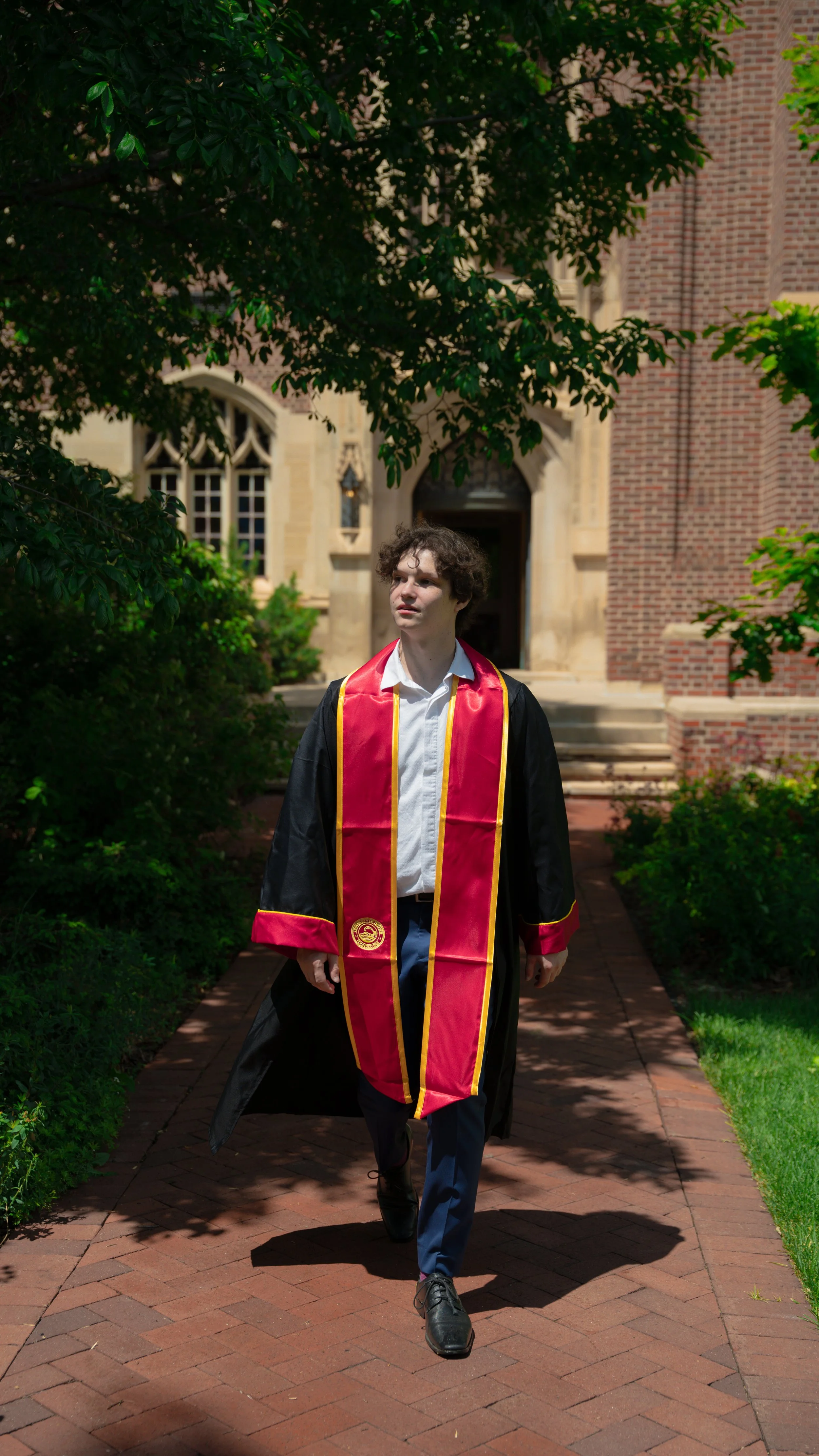 A young man in graduation gown and stole walking on brick pathway outside of a college building with trees and bushes.