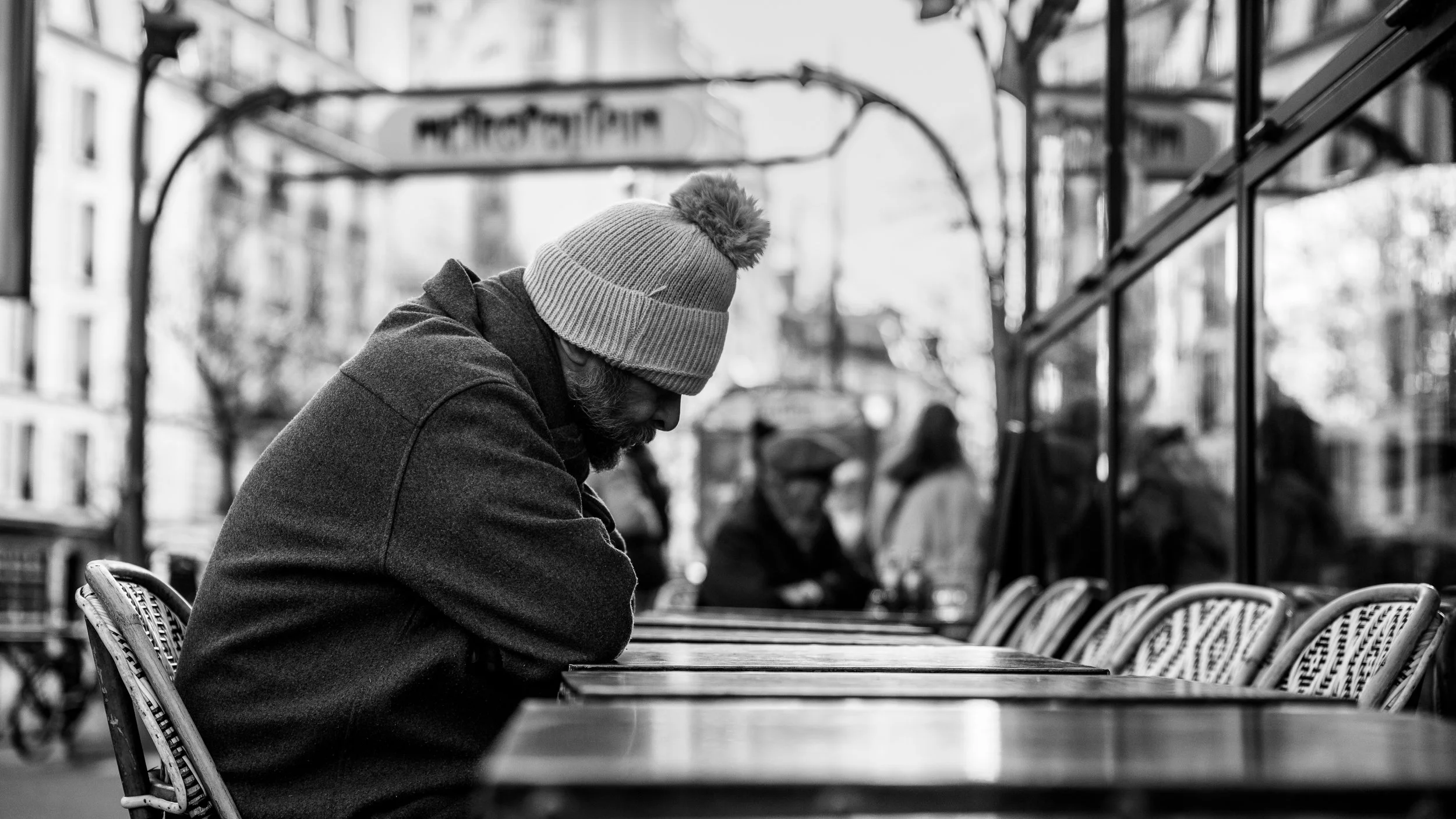 A man with a beard wearing a wool hat and dark coat sitting outdoors at a table at a cafe, with other people in the background, reflected in a window, in black and white.