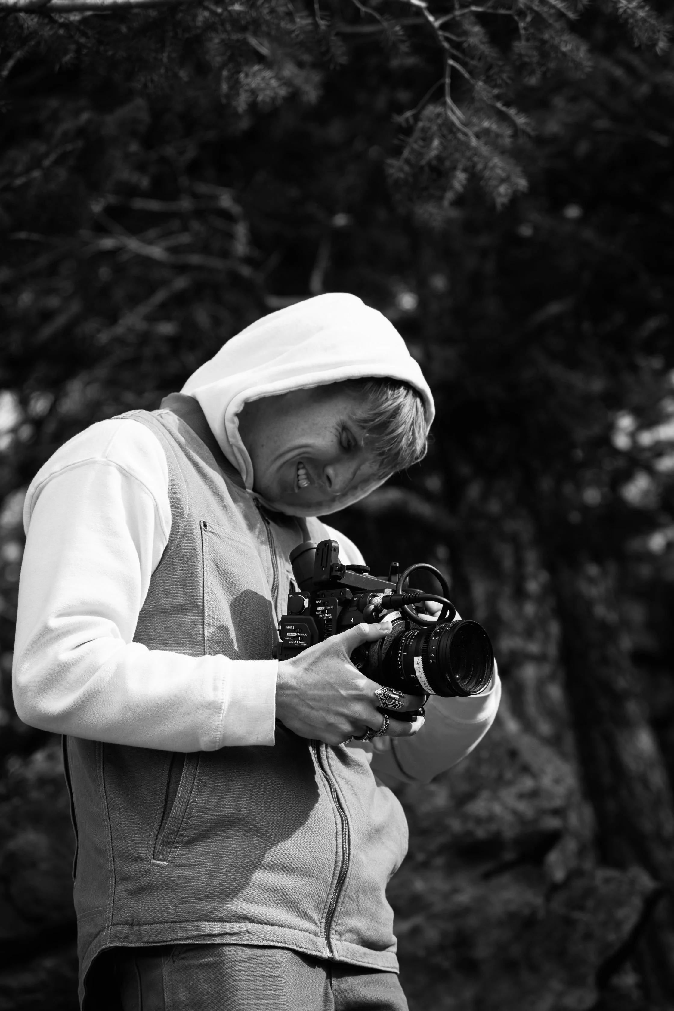 A person in a hoodie and jacket smiling while holding a professional video camera outdoors.