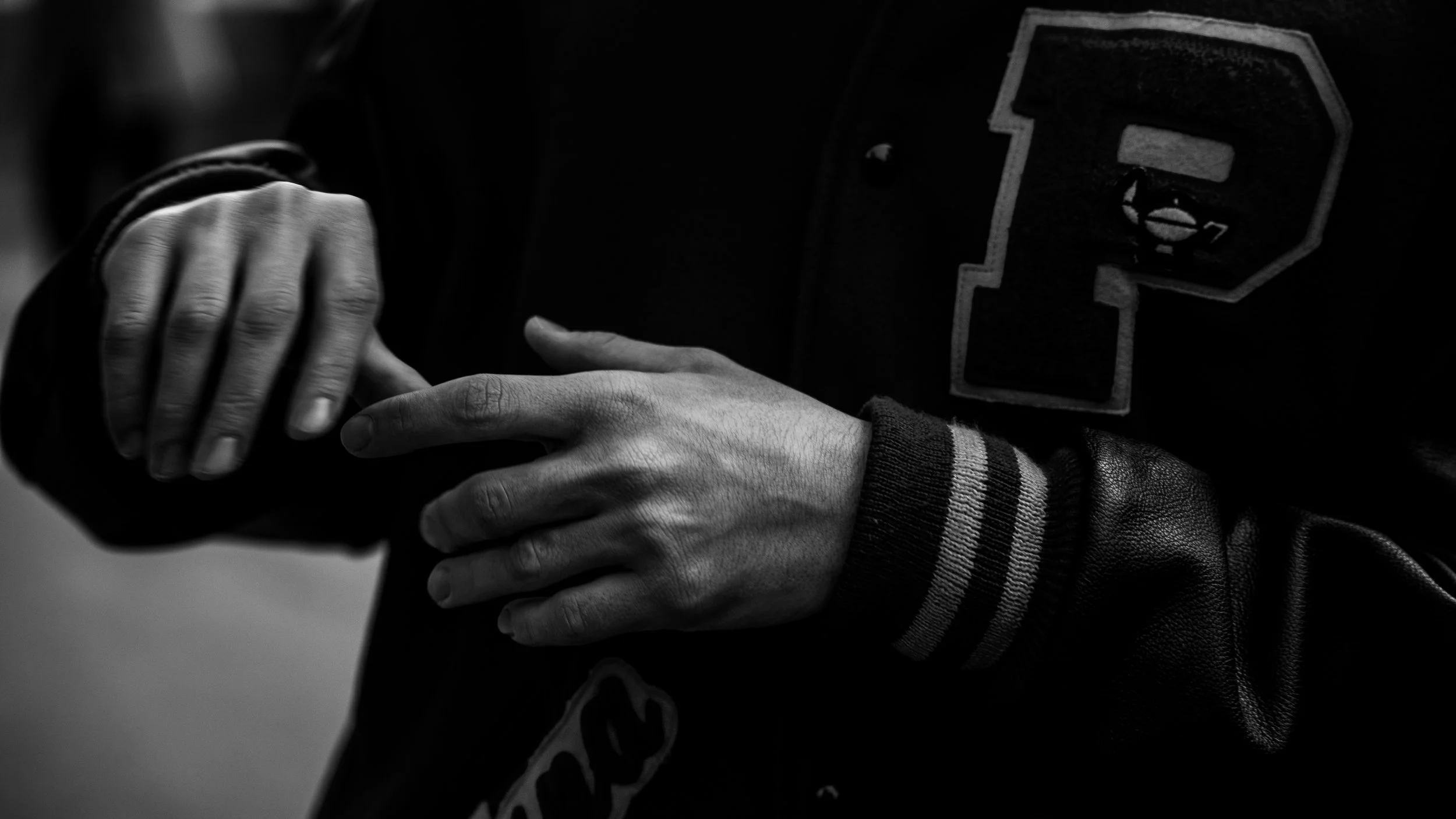 Close-up of a person's hands with fingers interlocked, wearing a black varsity jacket with striped cuffs and a large letter patch.
