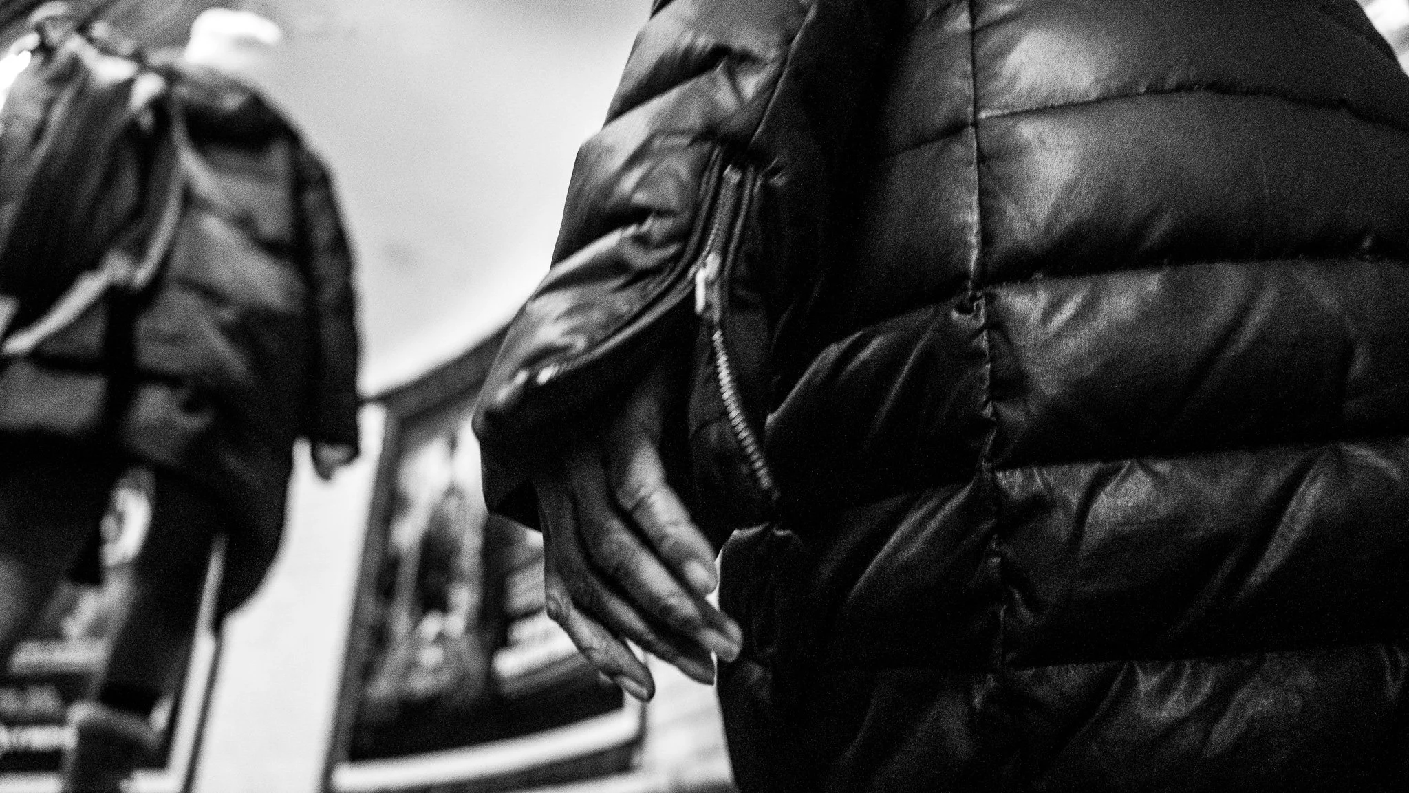 Close-up of a person's hand resting on their hip, wearing a shiny black puffer jacket, with a blurred person carrying a backpack in the background.