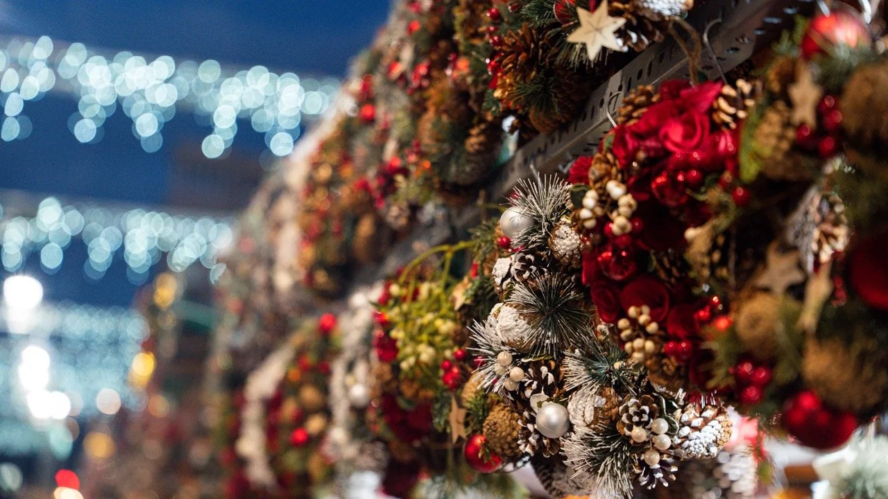 Close-up of Christmas decorations with red, white, and gold ornaments, pinecones, berries, and stars, with blurred Christmas lights in the background.