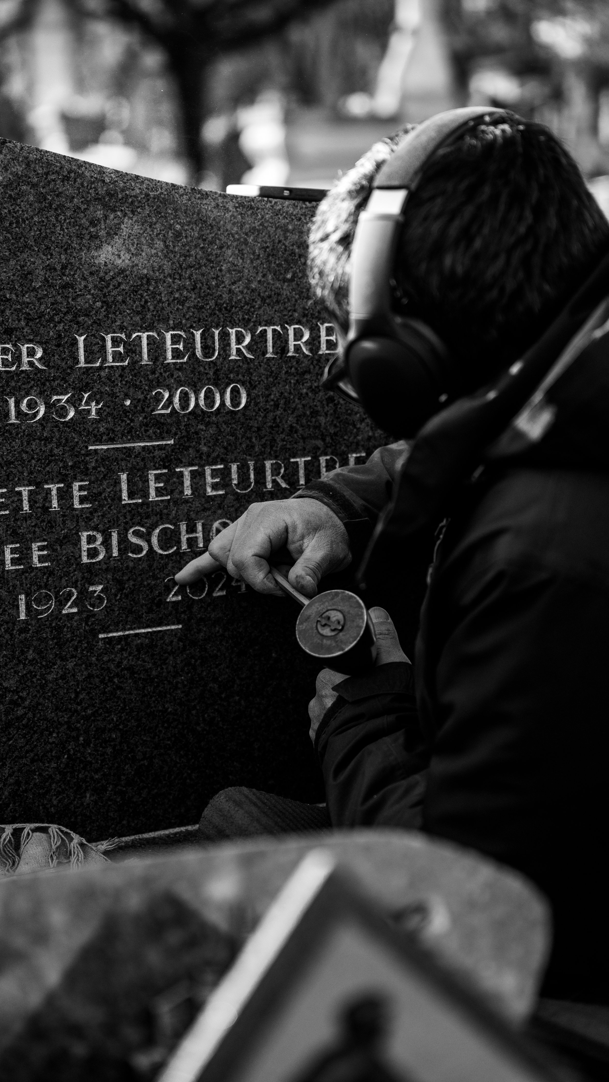 A person wearing headphones and a jacket is engraving a name on a gravestone with a small chisel and hammer. The gravestone has the names 'Peter Leteurtre' and 'Lettie Leteurtre' along with their birth and death years.